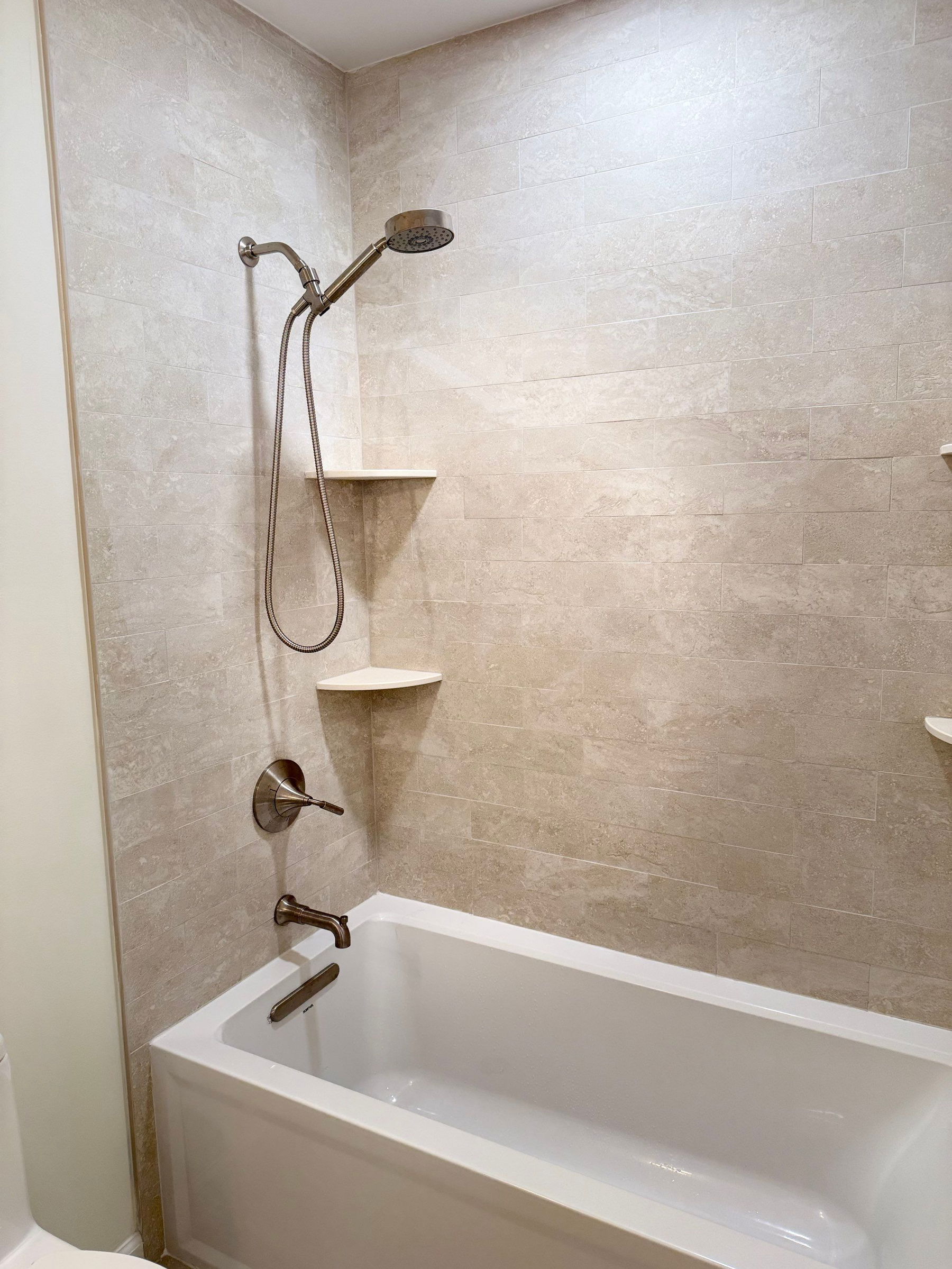 Bathroom shower alcove with beige linear tile, white built-in shelves, bronze rainfall showerhead with handheld wand, and white soaking tub below.