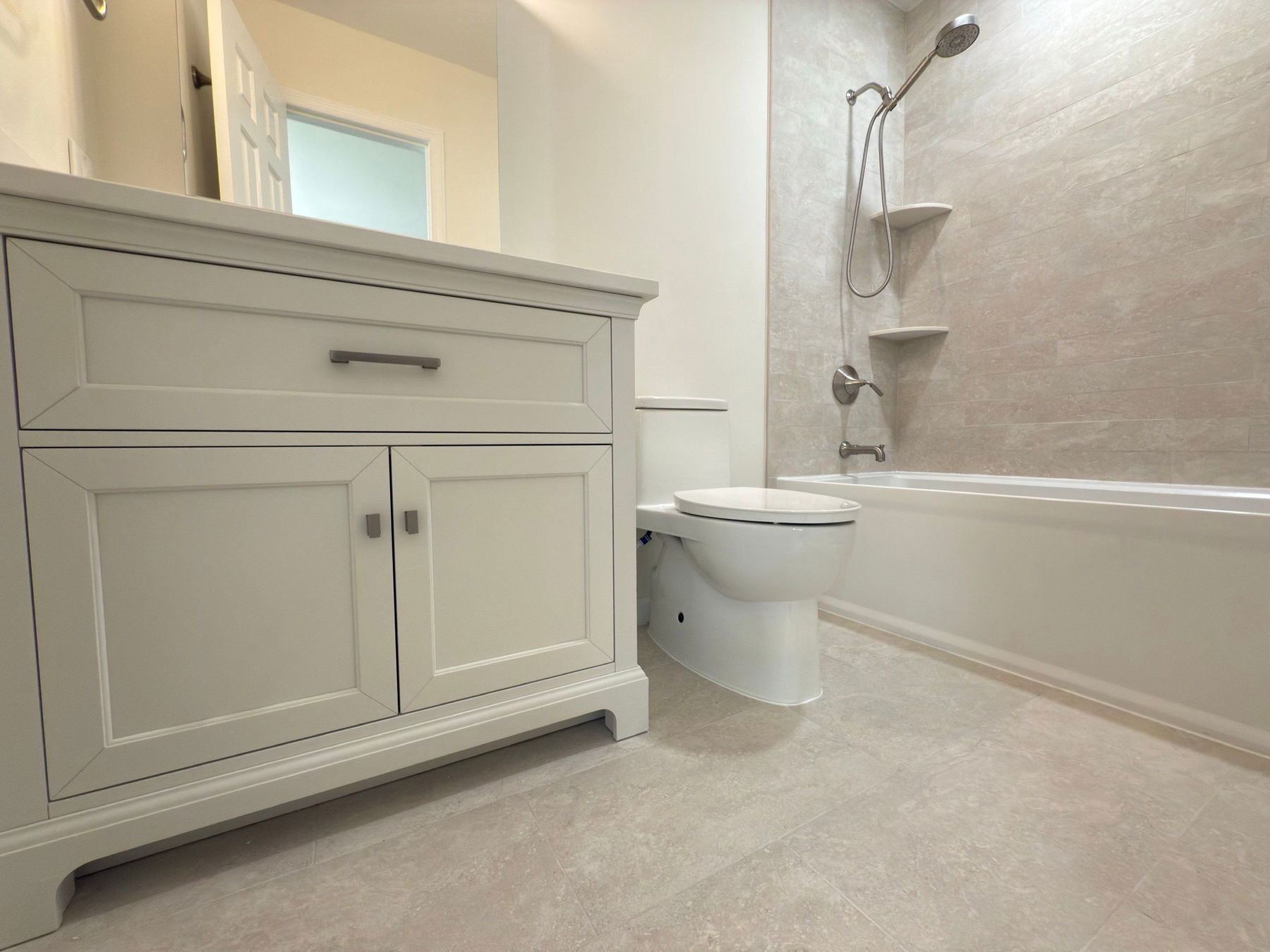 Light gray vanity with two cabinet doors and upper drawer, framed mirror, white toilet, bathtub with gray tile surround, chrome shower fixtures, and built-in shelving.