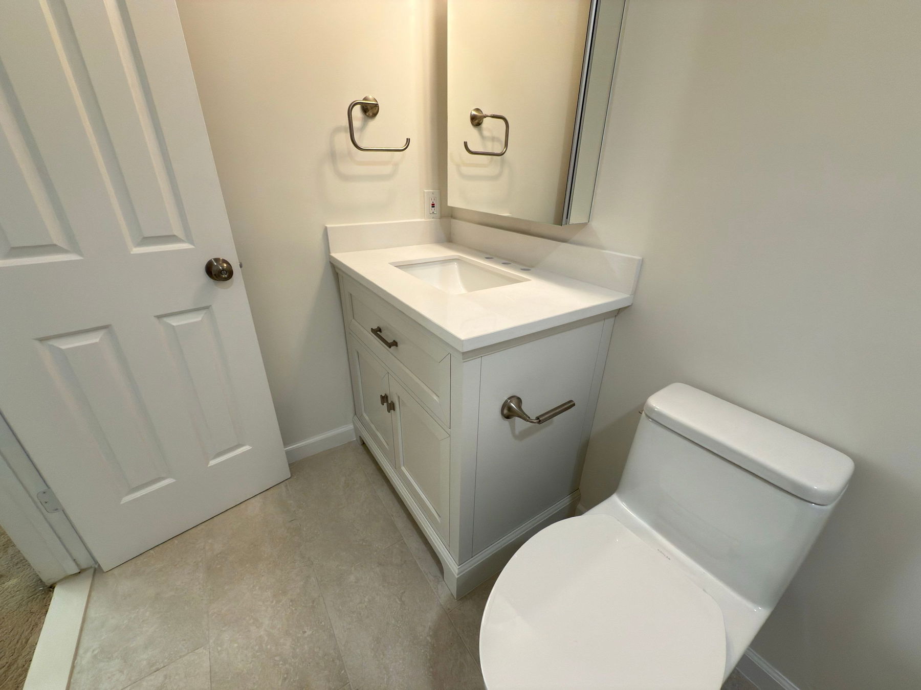 Compact bathroom with white vanity cabinet, white quartz countertop with integrated sink, brushed nickel hardware and towel rings, white toilet, beige tile flooring, and white six-panel door.