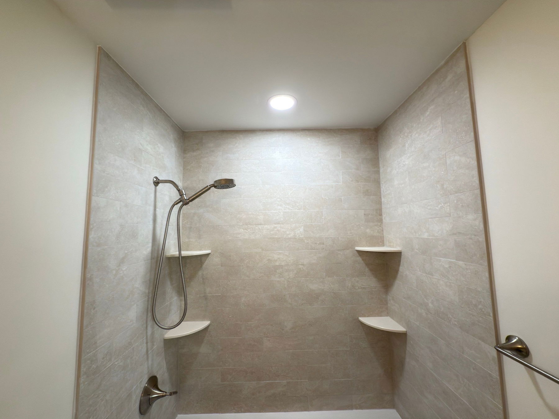 Walk-in shower with light gray tile walls, bronze fixtures, dual recessed shelving, and recessed ceiling light with frameless glass enclosure.
