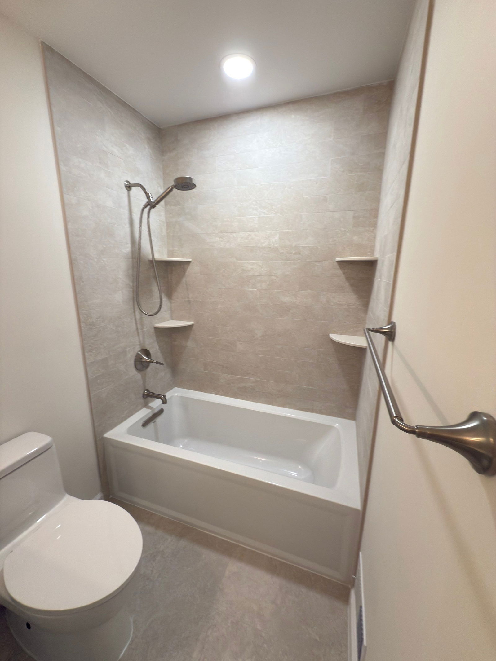 Bathroom with white alcove tub, beige tile surround with recessed shelves, chrome fixtures, grab bar, and recessed ceiling light.