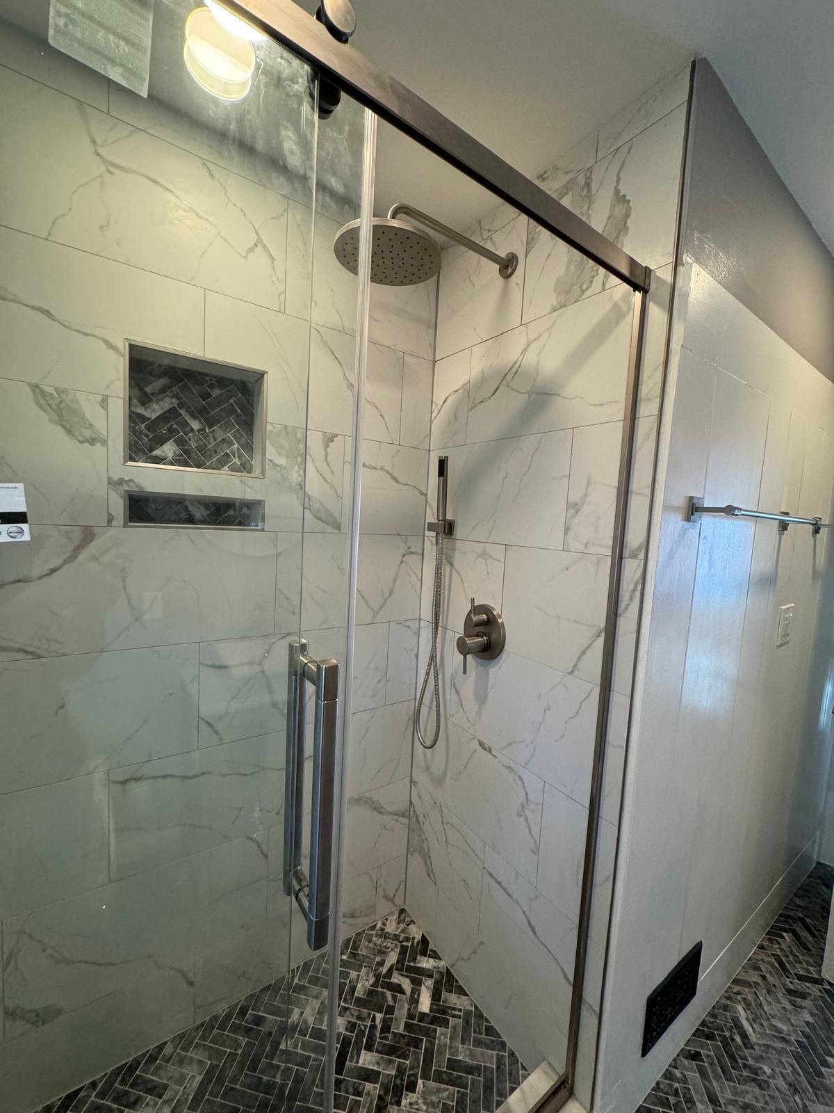 Luxury bathroom shower in Belle Mead featuring large-format marble-look porcelain tiles with veining, frameless glass enclosure, stainless steel fixtures, rainfall showerhead, and herringbone-pattern dark tile floor.