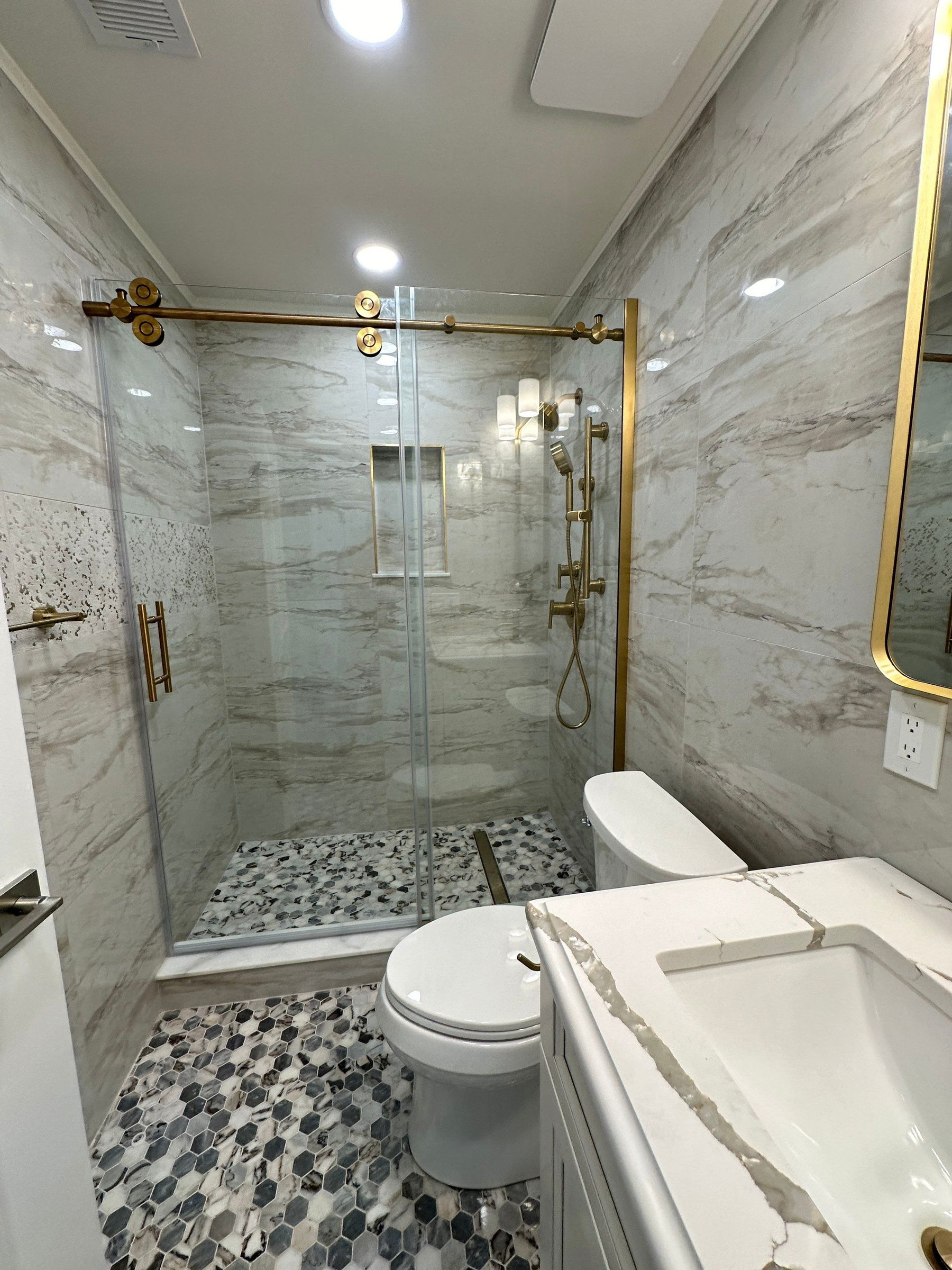 Marble-tiled bathroom with frameless glass walk-in shower, brass fixtures, hex mosaic floor, white vanity, and recessed lighting.