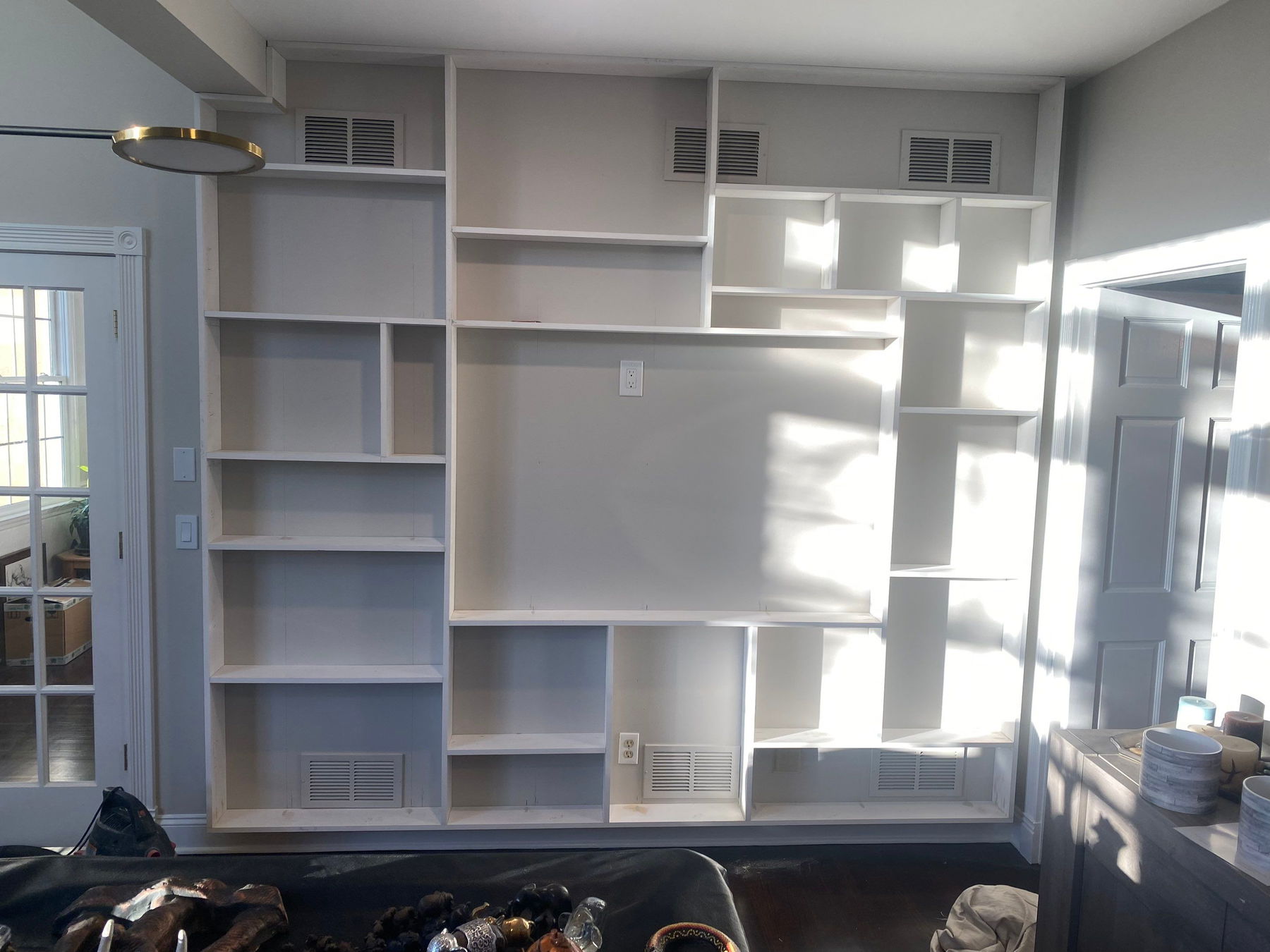 Built-in wall shelving unit with white painted wood frame, asymmetrical open shelving, integrated ventilation louvers, and brass wall sconce, flooded with natural sunlight.