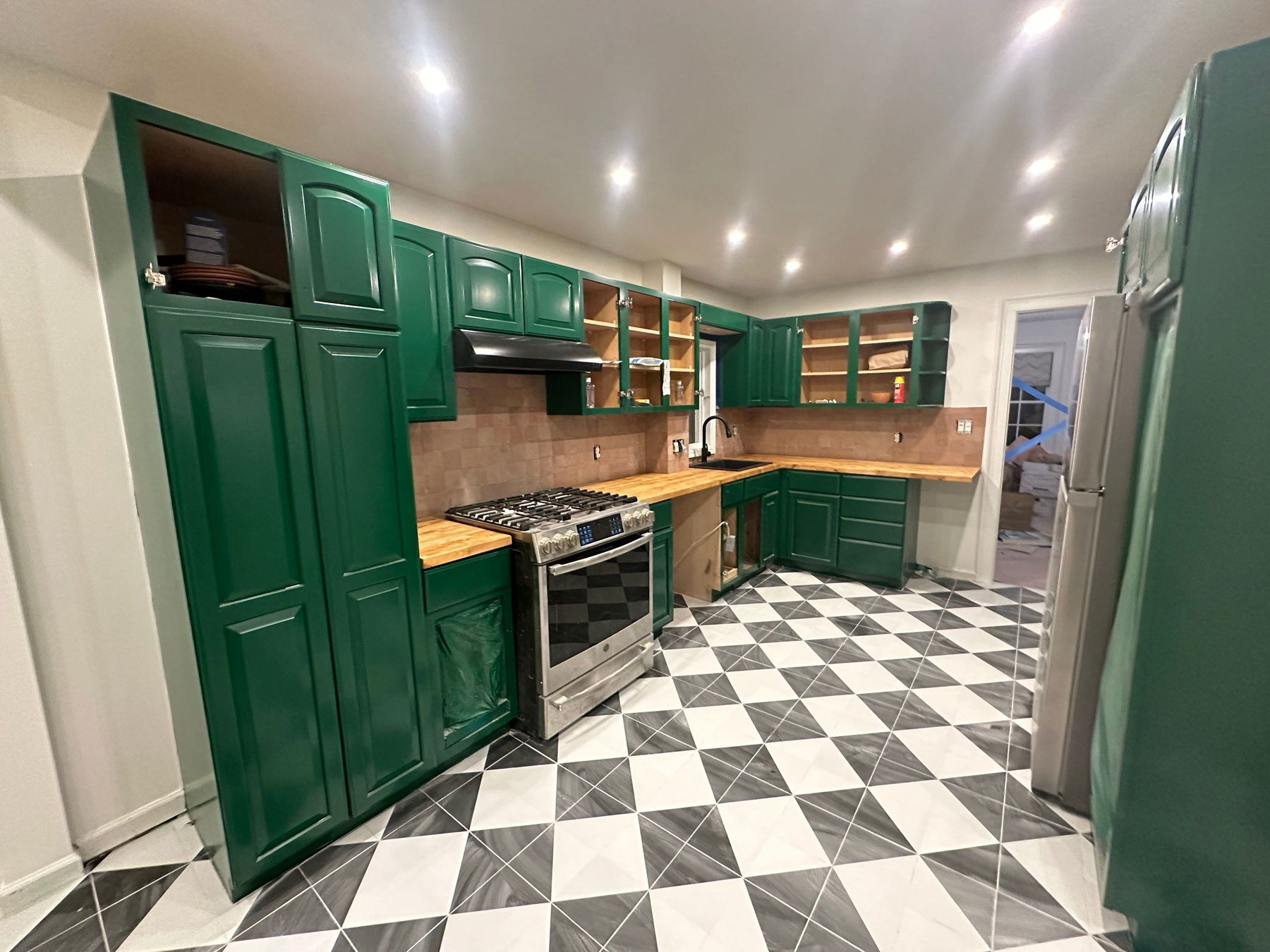 Modern kitchen with forest green shaker cabinets, butcher block countertops, tan tile backsplash, stainless steel gas range, open shelving, recessed ceiling lights, and black-and-white diamond-pattern floor tile.