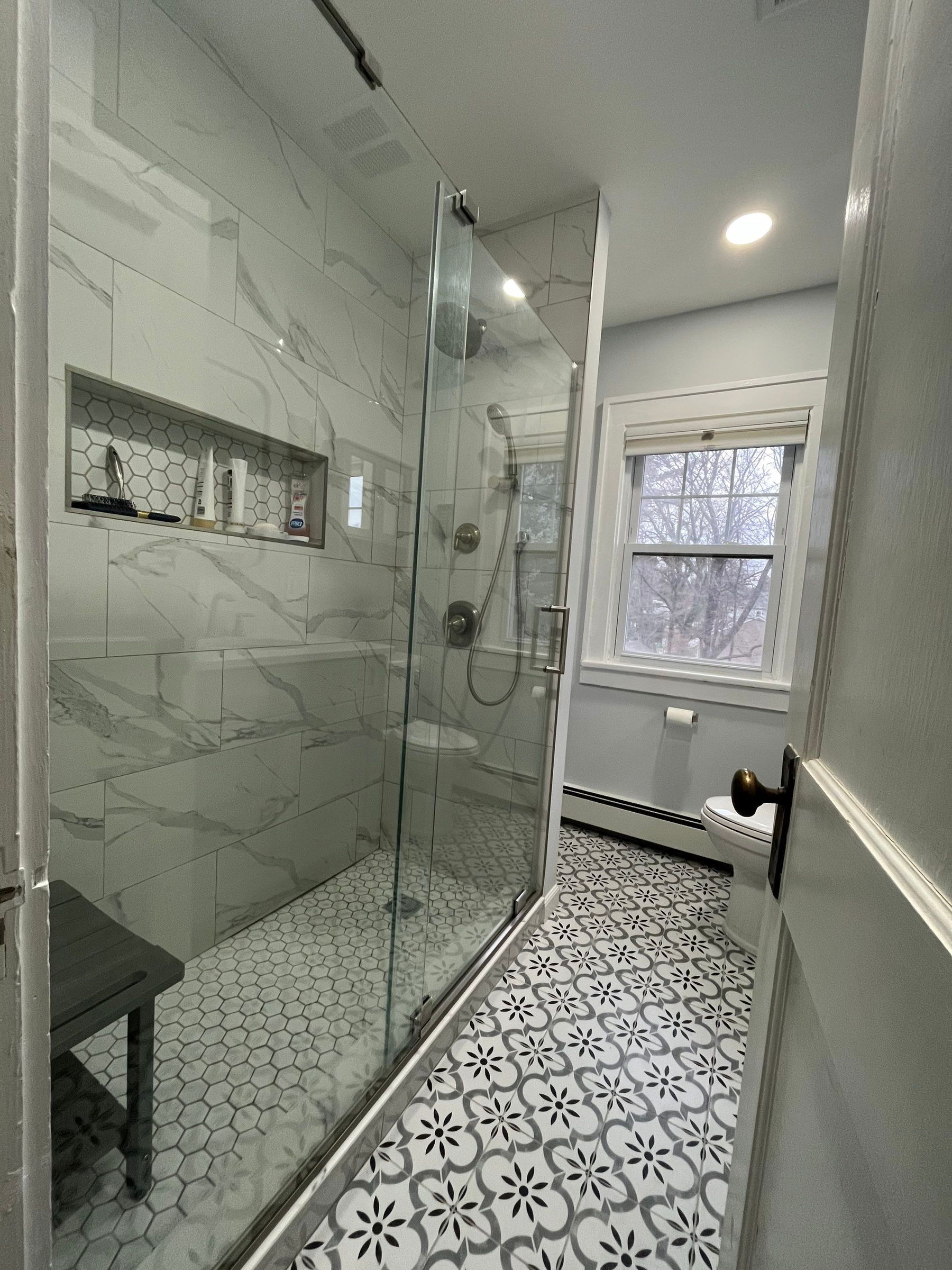 Modern bathroom with marble-look tile shower surround, glass enclosure, hexagonal floor tile, pedestal sink, and window with natural light.