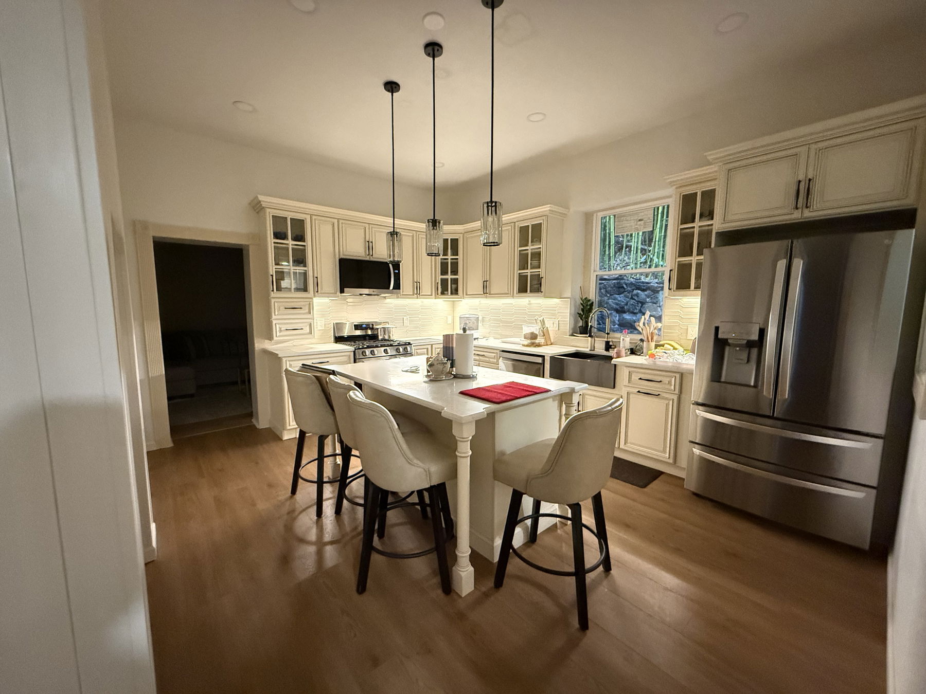 Cream cabinetry with glass doors, white subway tile backsplash, stainless steel appliances, white quartz island with turned legs, black pendant lights, and wood flooring.