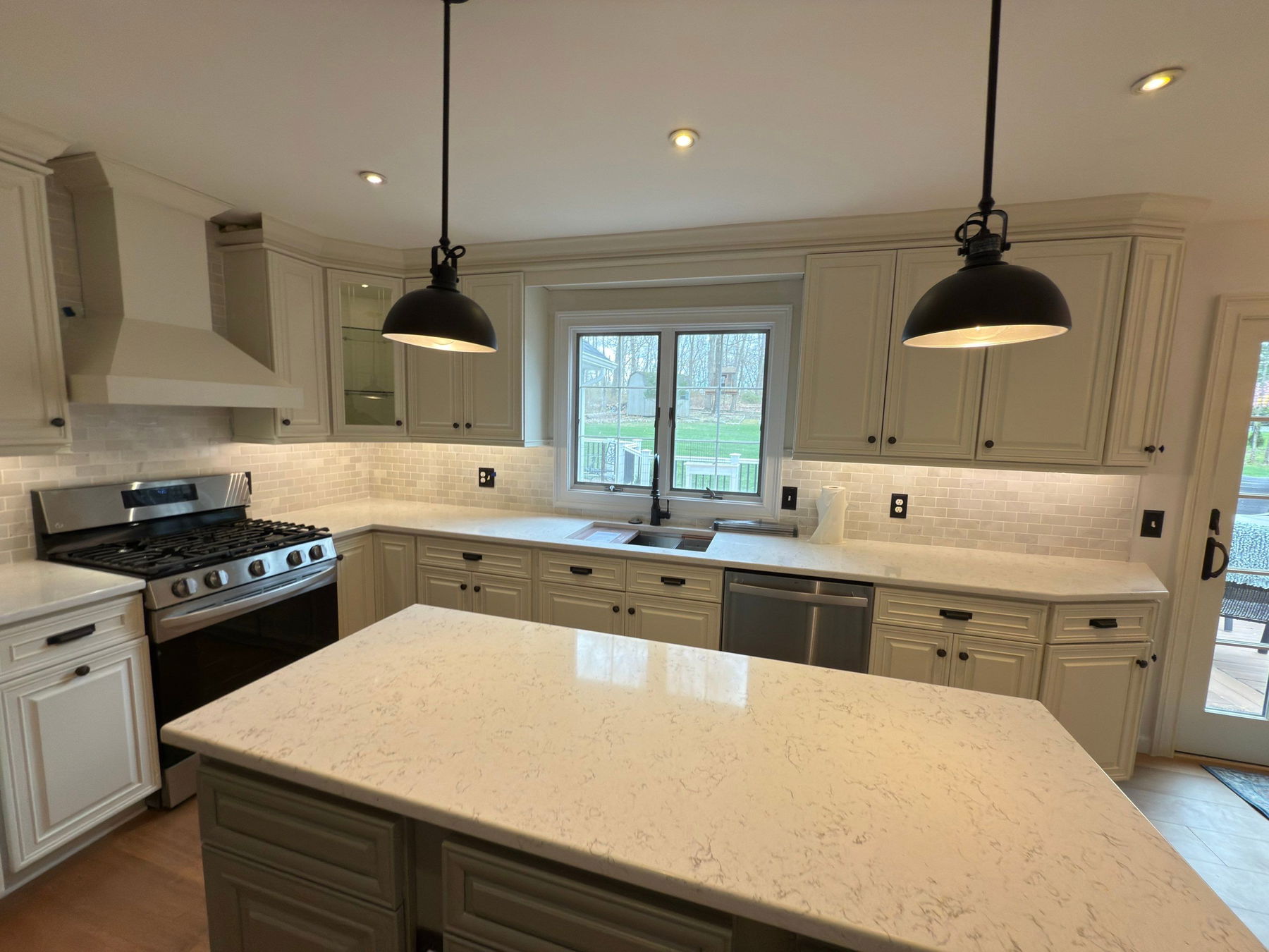 Cream-colored shaker cabinetry with marble-look quartz countertops, stainless steel gas range, white subway tile backsplash, black pendant lights, and French doors overlooking a backyard.