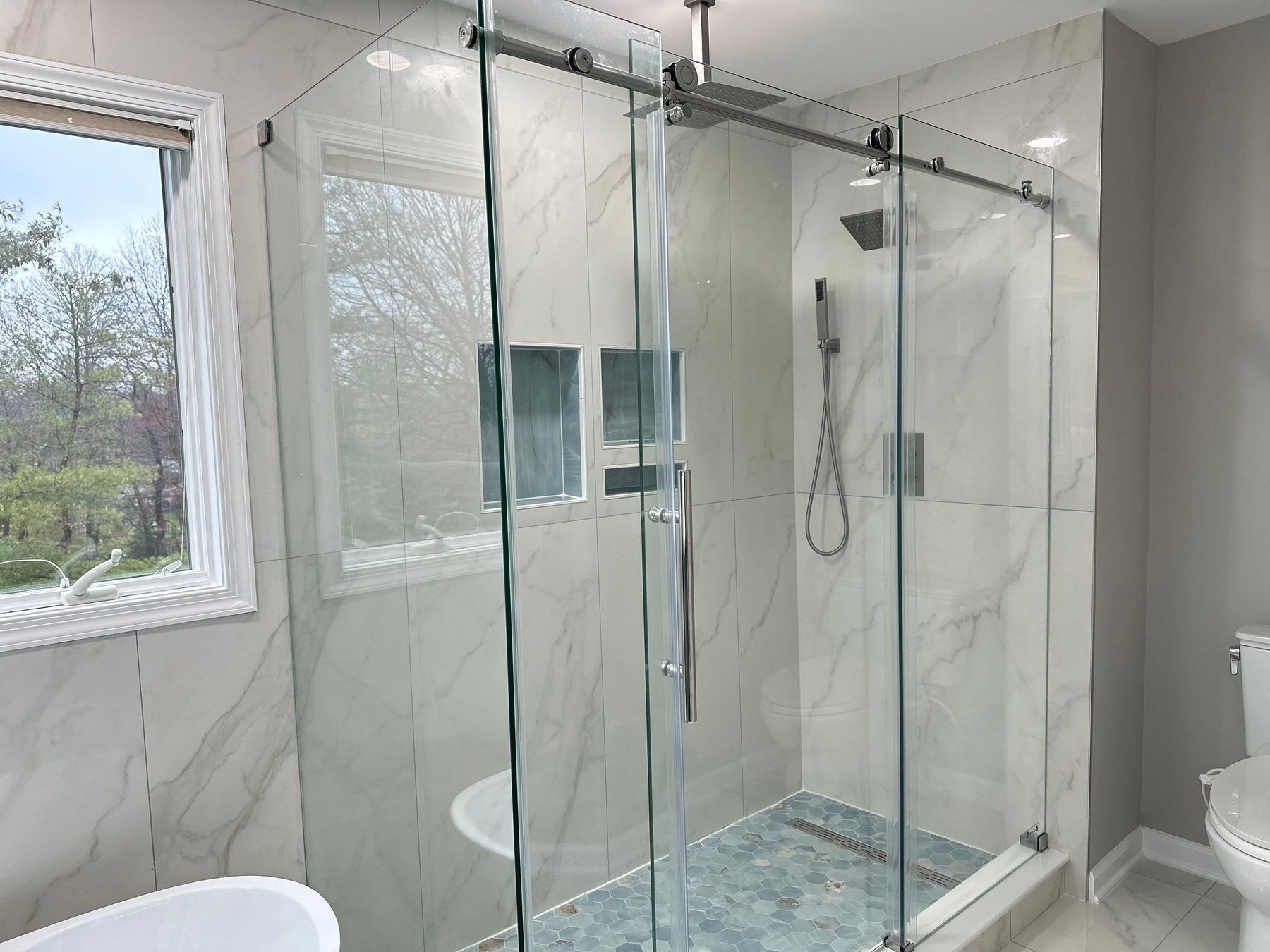 Spacious shower enclosure with frameless glass sliding doors, marble-look tile walls, rainfall showerhead, hand shower, hexagonal floor tiles, and large window with view of trees.