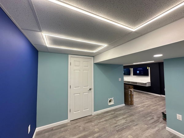 Finished basement with teal and navy accent walls, white six-panel door, linear LED ceiling fixtures, gray wood-look flooring, and built-in entertainment area with black cabinetry and mounted TV.