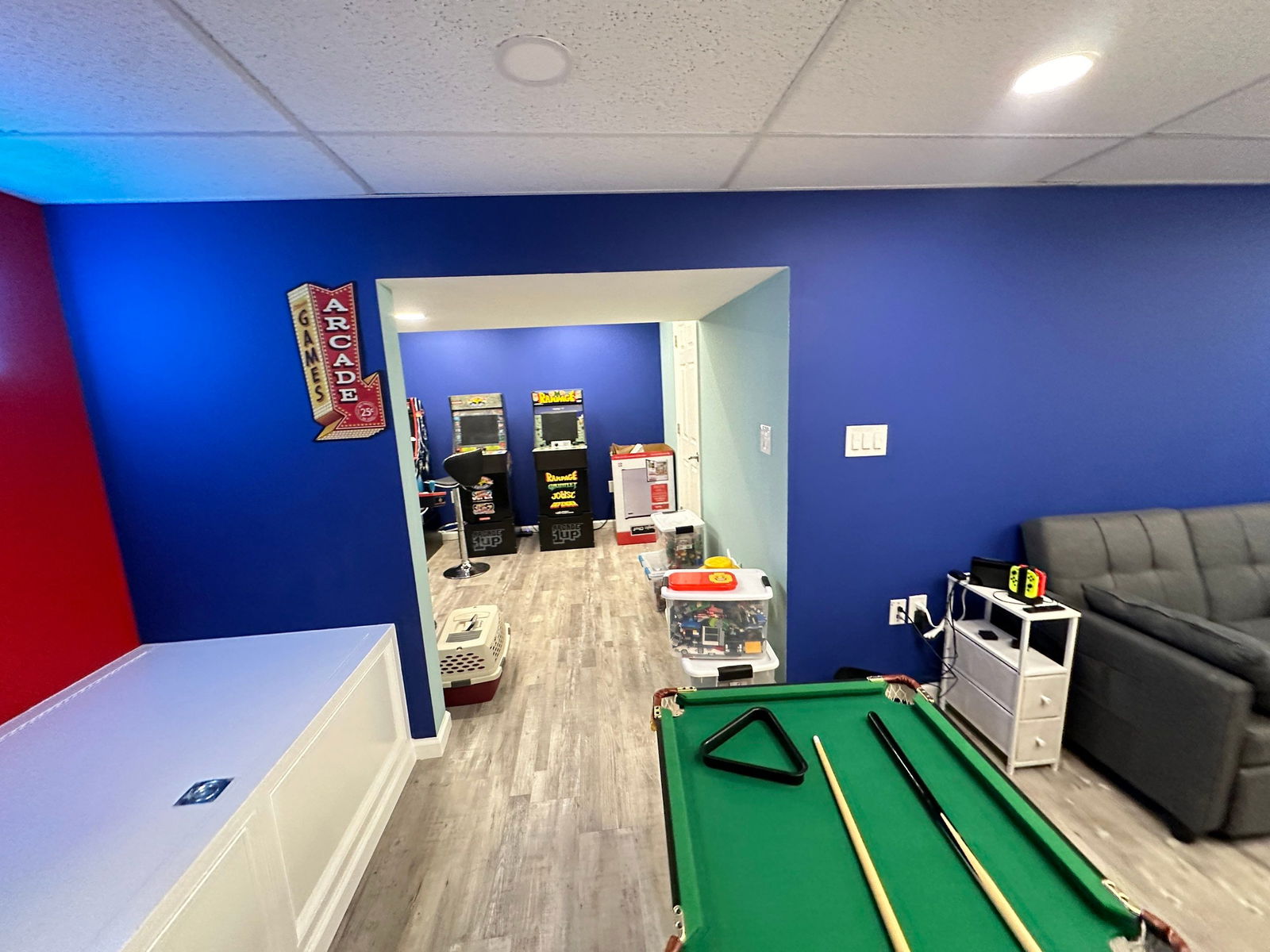 Finished basement recreation room with bright blue walls, wood-look plank flooring, pool table, air hockey table, arcade machines, and vintage neon sign.