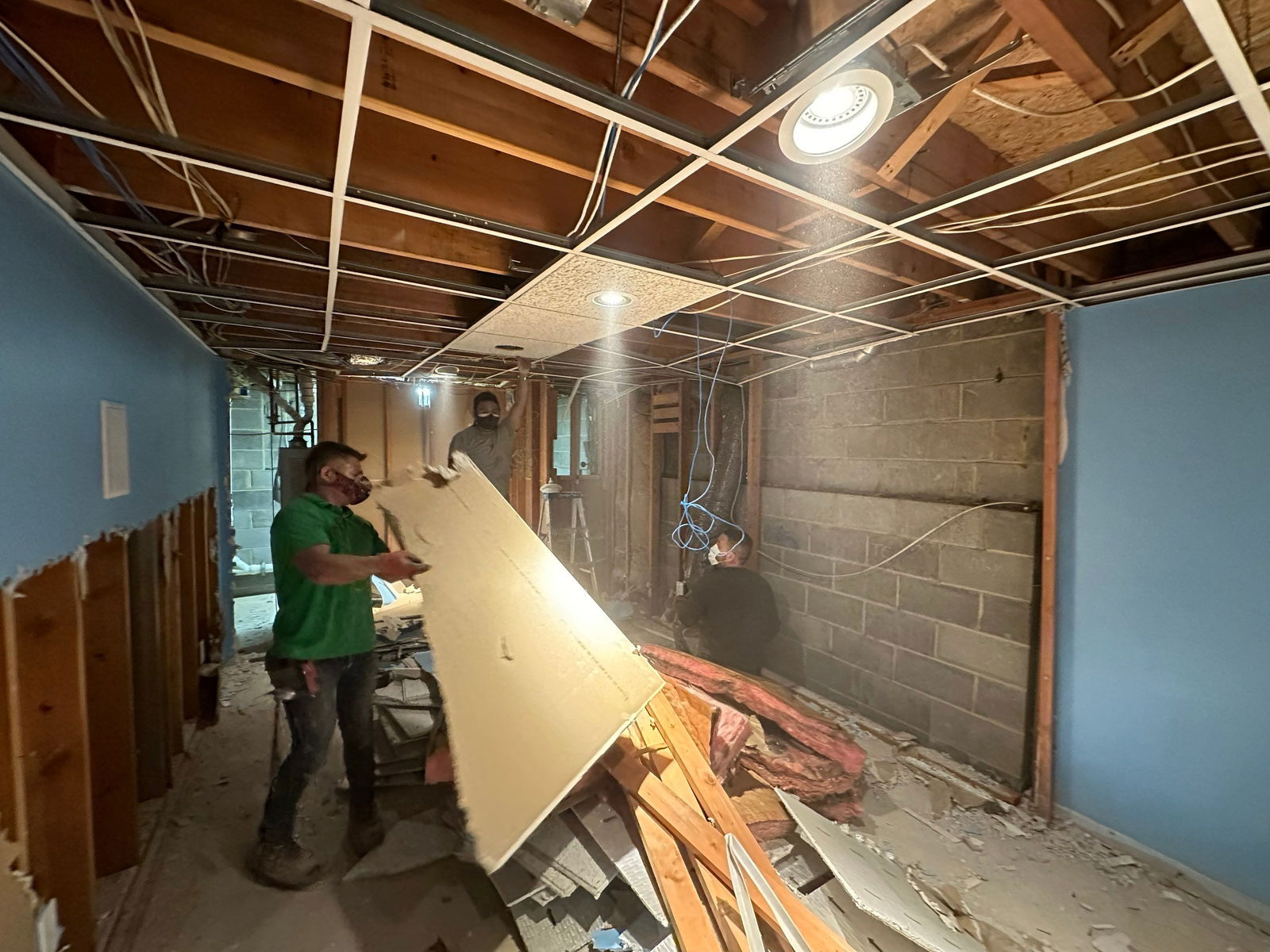 Basement renovation in progress with exposed framing, suspended ceiling grid, recessed lighting, blue-painted walls, and drywall sheets being installed by workers.