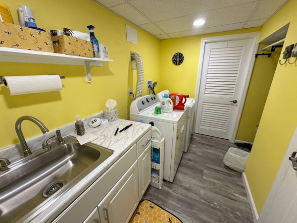 Bright yellow laundry room with white cabinetry, stainless steel sink, marble countertop, white louvered doors, gray wood-look flooring, and open shelving.