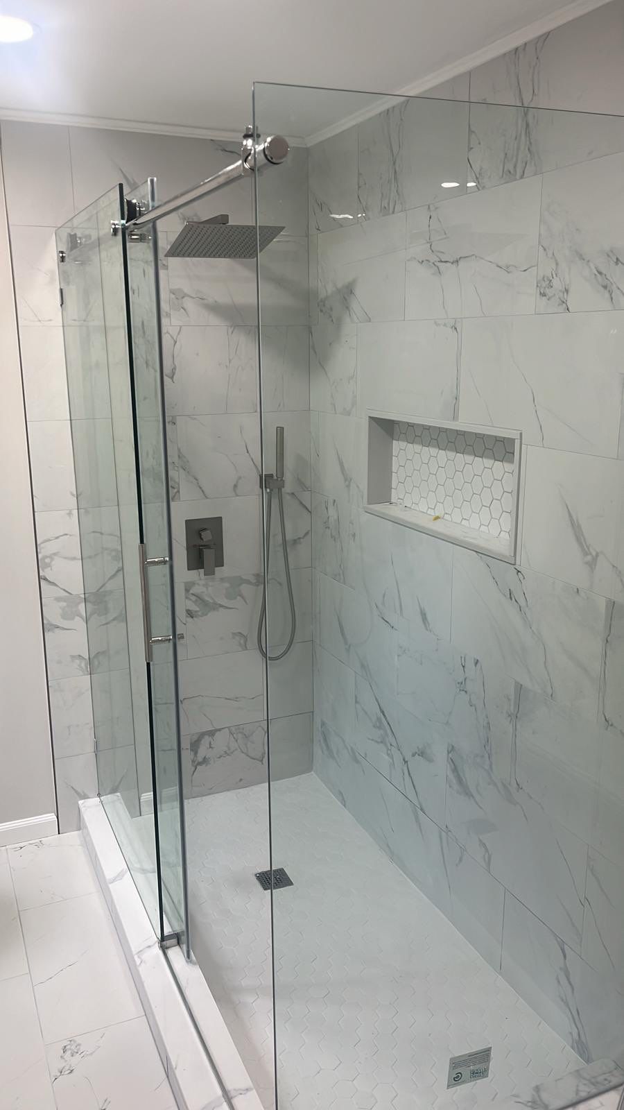 Walk-in shower with frameless glass enclosure, gray and white marble-look large-format tile walls, chrome square rainfall showerhead, matte black fixtures, and built-in niche with white hexagon mosaic tile accent.