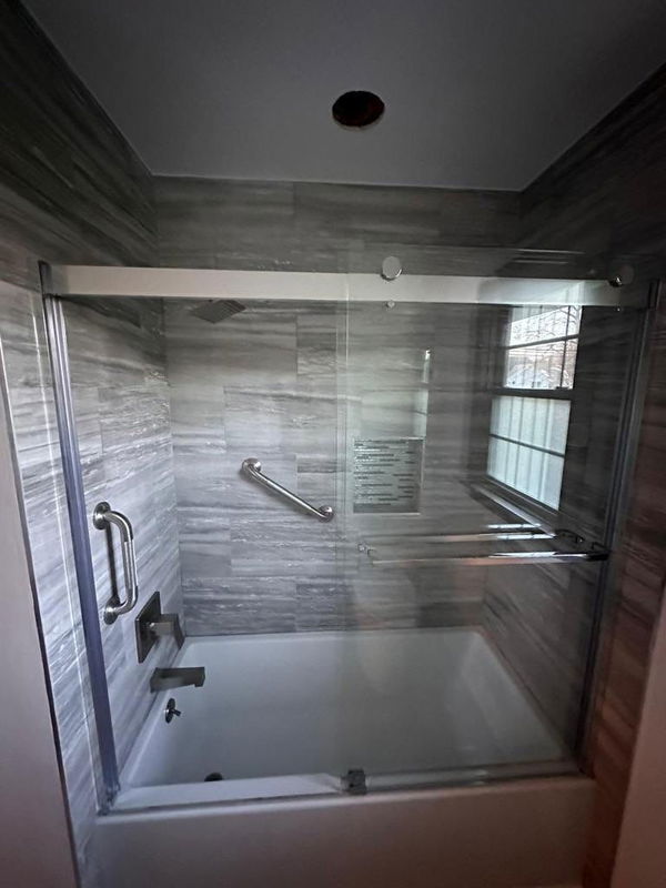 Frameless glass shower enclosure with gray linear tile walls, chrome grab bar and handles, white soaker tub, recessed ceiling light, and chrome trim.