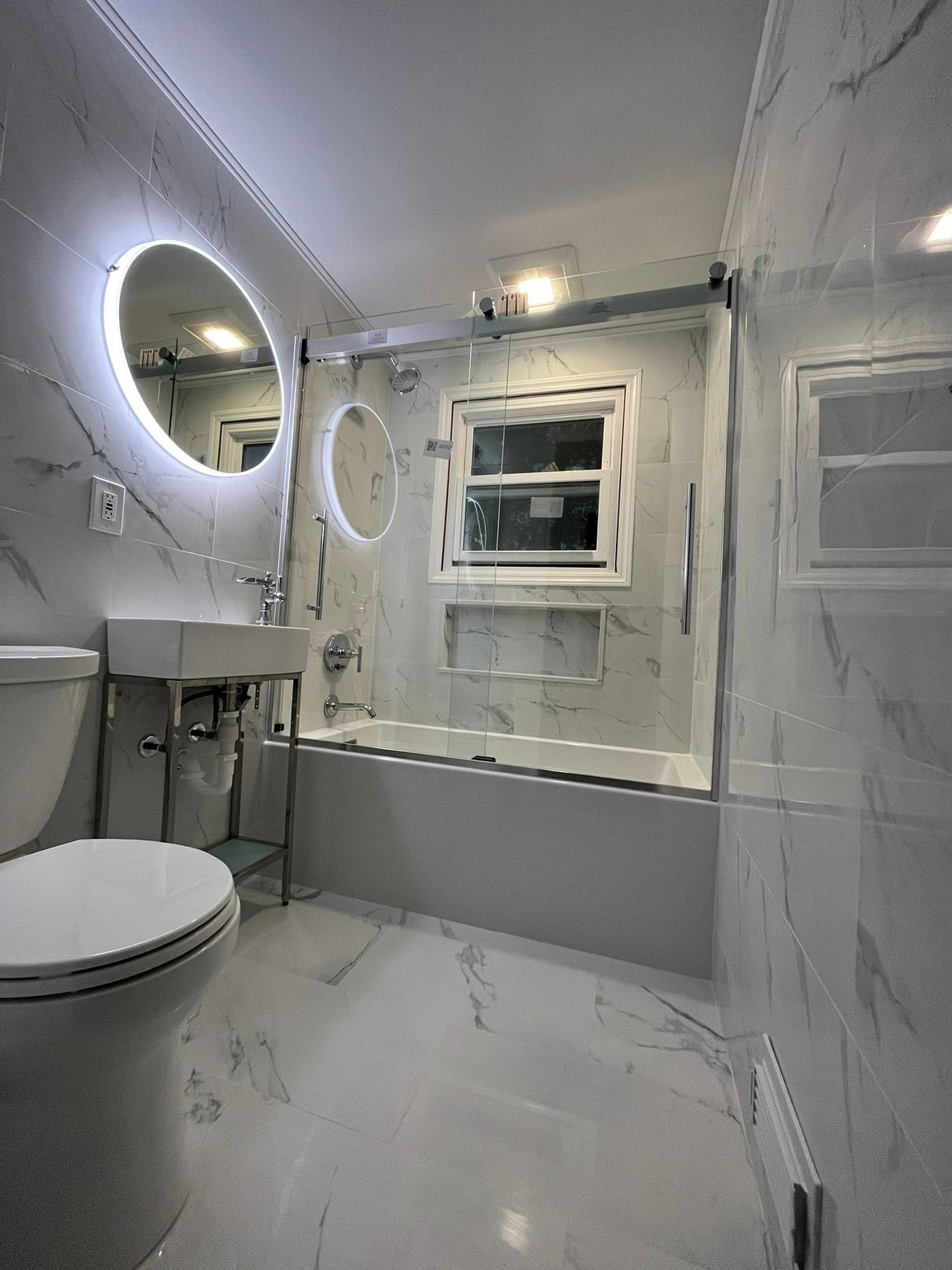 Modern bathroom with white marble-look tile walls and floor, frameless glass shower enclosure with chrome trim, floating white sink vanity, illuminated round mirror, and toilet.