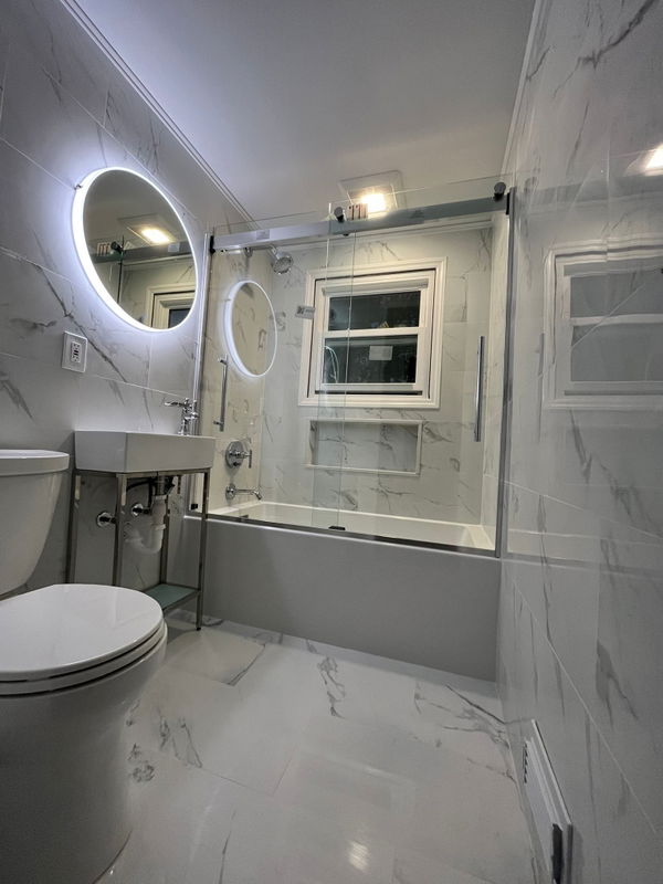 Modern bathroom with white marble-look tile walls and floor, frameless glass shower enclosure with chrome trim, floating white sink vanity, illuminated round mirror, and toilet.