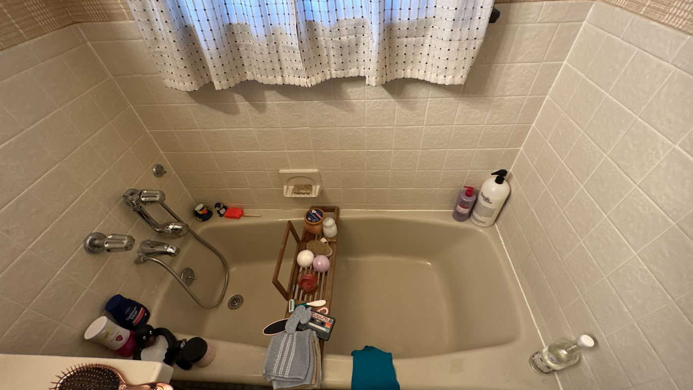 Beige tile bathroom with soaking tub, chrome fixtures on left wall, white perforated shower curtain, wooden caddy with bath accessories, and various toiletries on tub ledge.