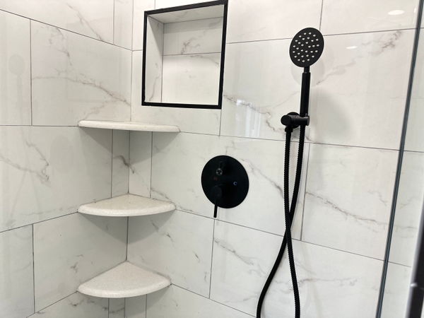 Walk-in shower with white marble-look large-format tiles, black matte showerhead and valve, built-in corner shelving, and recessed niche with black frame.