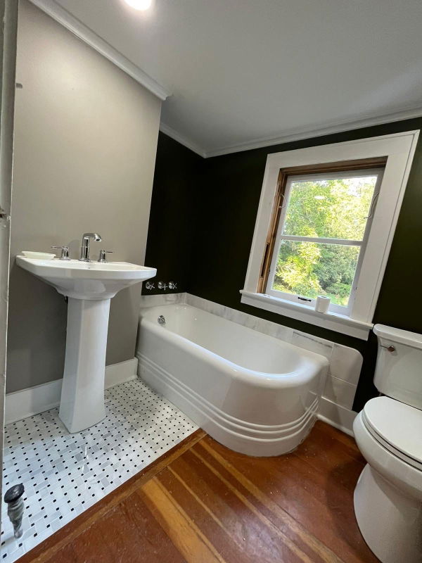Bathroom with white pedestal sink, freestanding soaking tub, black accent wall, white subway tile surround, wood flooring, polished chrome fixtures, and large window with natural light.