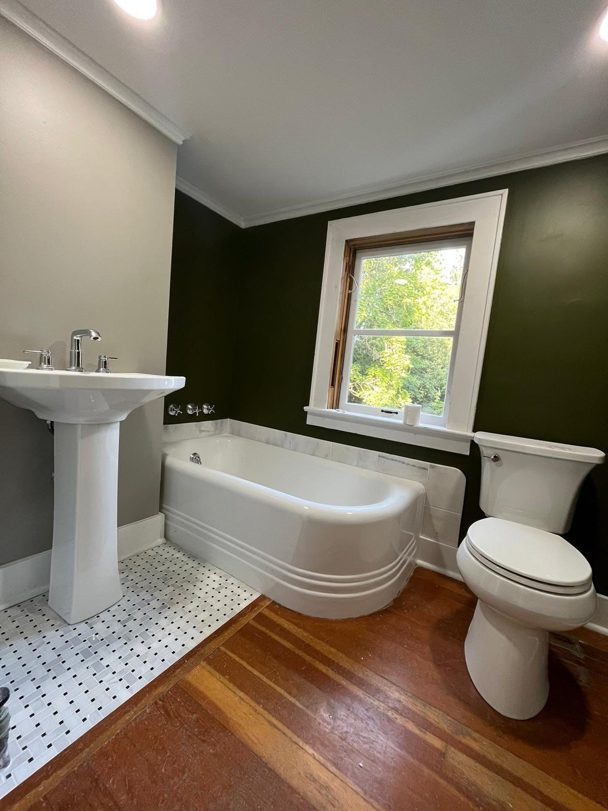 Master bathroom with white pedestal sink, curved white soaking tub, white toilet, dark green accent wall, white subway tile surround, polka-dot floor tile, wood flooring, recessed lighting, and wood-trimmed window with green landscape view.
