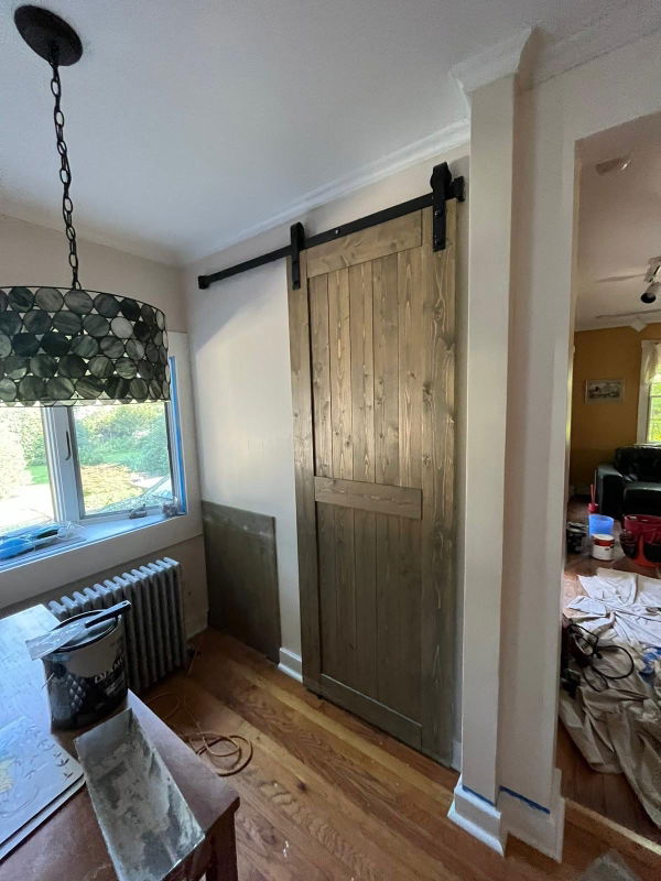 Light wood barn door with black hardware sliding across white wall opening, cream-colored ceiling with crown molding, hardwood floor, and window with geometric pendant light.