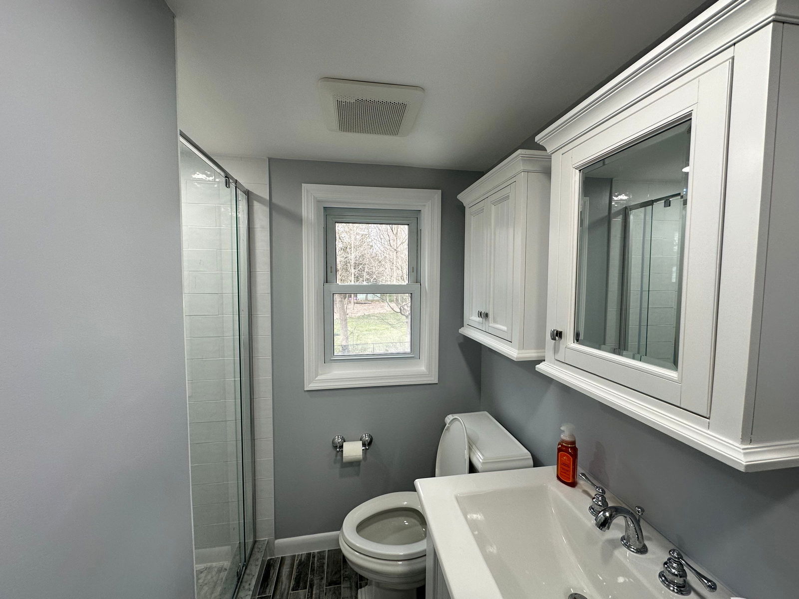 Gray bathroom with white pedestal sink, chrome faucet, white toilet, glass-enclosed shower with subway tile, large medicine cabinet with mirror, window with white trim overlooking landscaping, and ceiling exhaust vent.