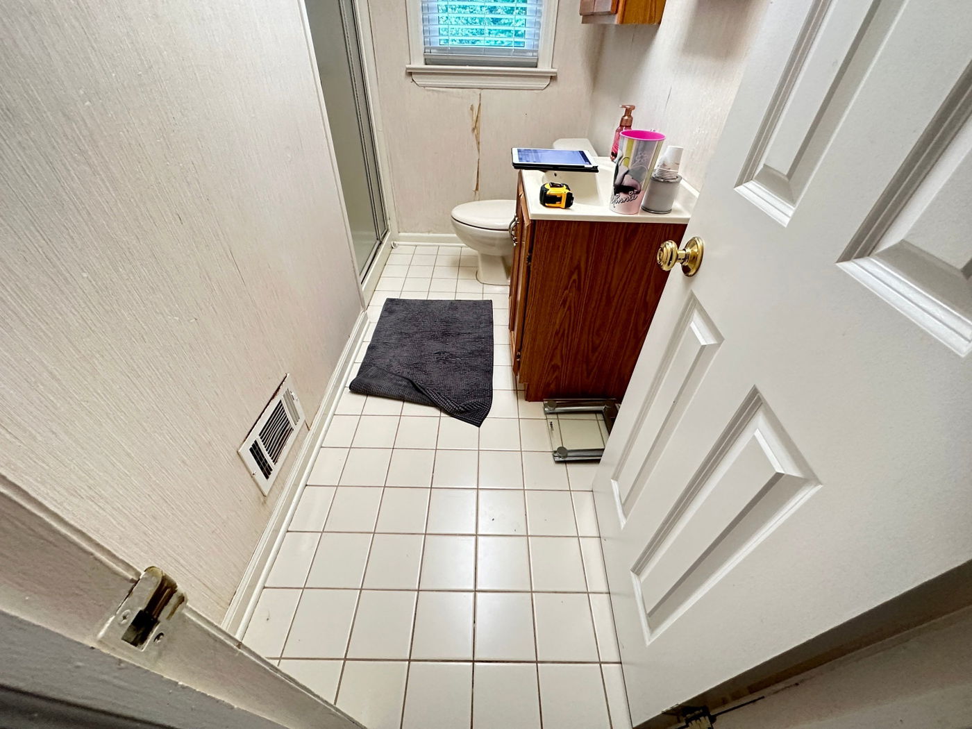 Small bathroom with white ceramic tile floor, cream textured walls, white toilet, wood-grain vanity with brass hardware, small window, and white trim throughout.
