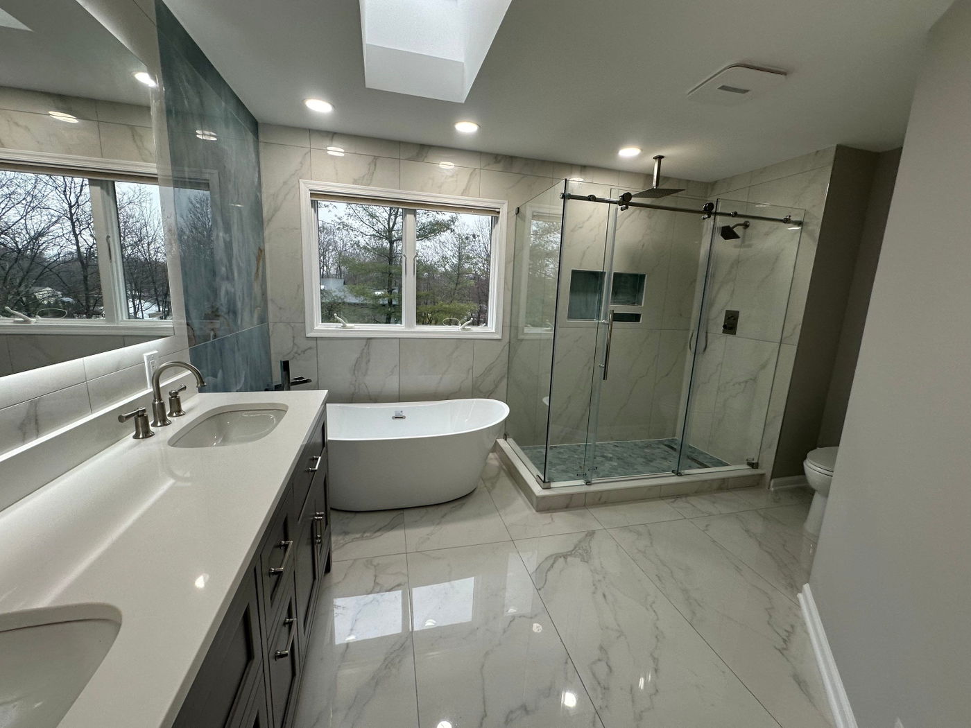 Spa-style bathroom with white marble flooring, marble-tiled walls, freestanding soaking tub, dual vanity with dark cabinetry, frameless glass shower enclosure with black fixtures, and skylights.