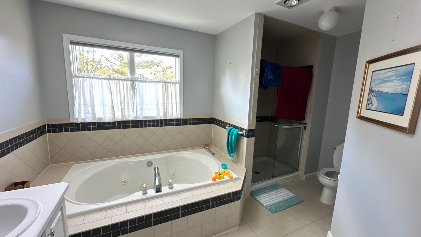 Master bathroom with white jetted soaking tub, black and cream tile surround, separate walk-in shower with glass enclosure, toilet, and neutral tile flooring.