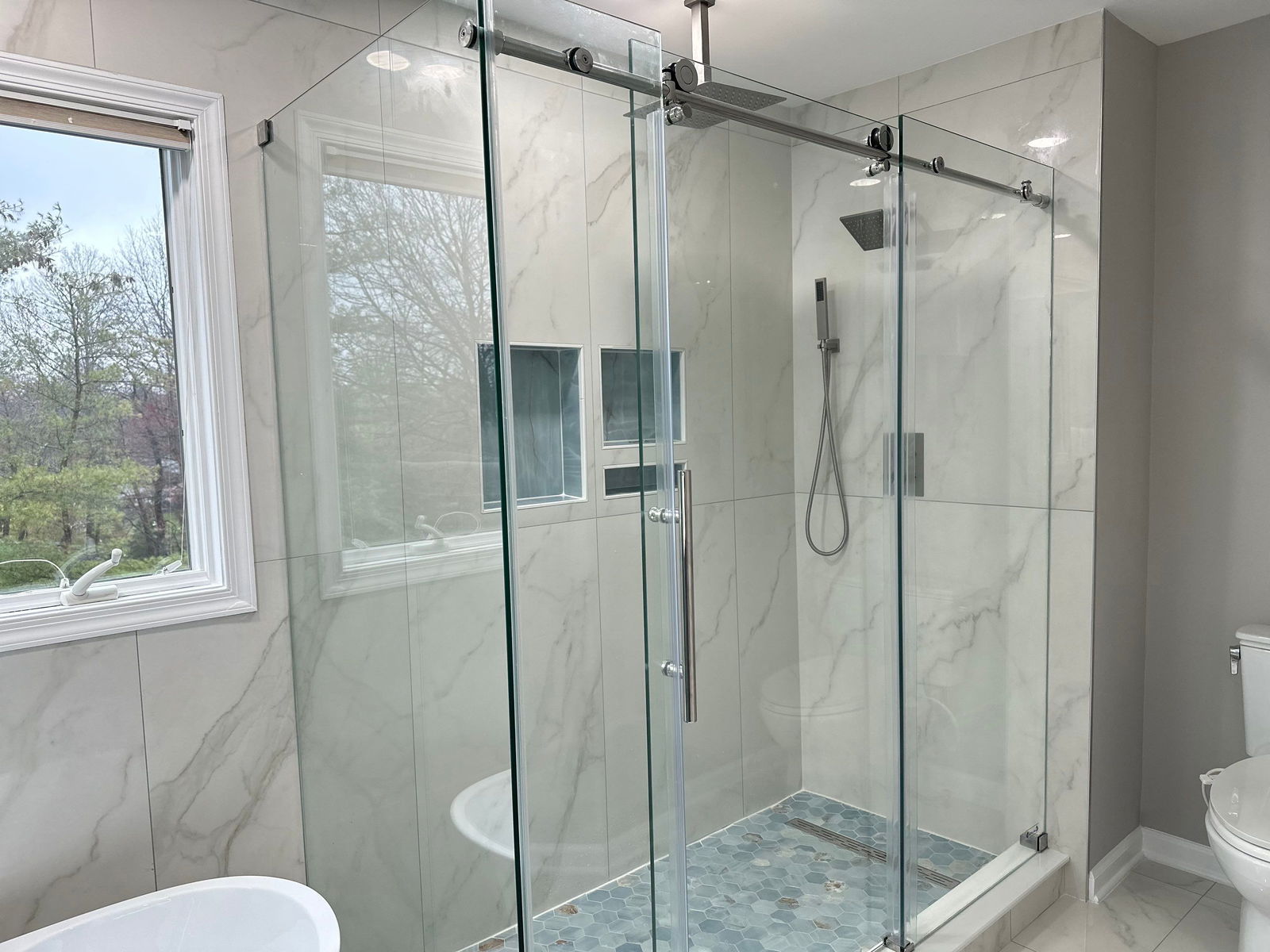 Spacious shower enclosure with frameless glass sliding doors, marble-look tile walls, rainfall showerhead, hand shower, hexagonal floor tiles, and large window with view of trees.
