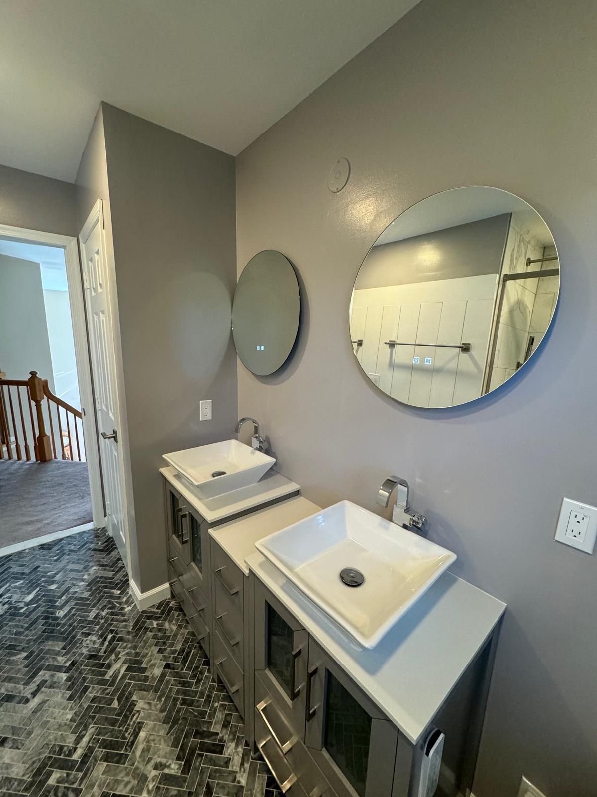 Belle Mead bathroom remodeling features dual white vessel sinks on gray cabinetry, round mirrors, herringbone tile flooring, and soft green walls with chrome fixtures.