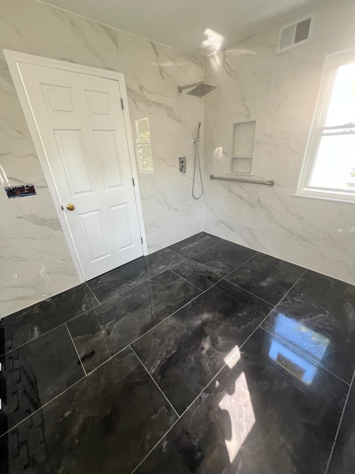Bathroom Remodeling in Bridgewater, NJ — Home Concepts Construction Spacious bathroom with white marble-look tile walls, black porcelain floor tiles, glass-enclosed shower with rainfall showerhead and hand rail, white six-panel door, and bright windows.