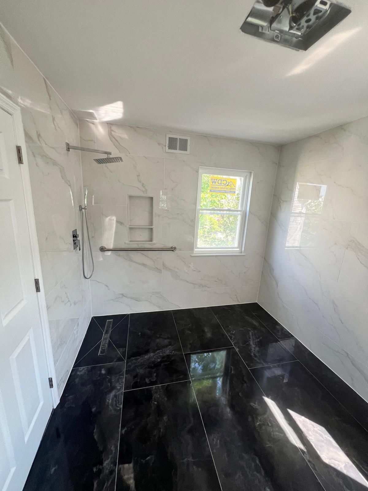 Spacious bathroom with white marble-veined tile walls, black polished floor tile, frameless glass shower enclosure with chrome fixtures, and two windows providing natural light.