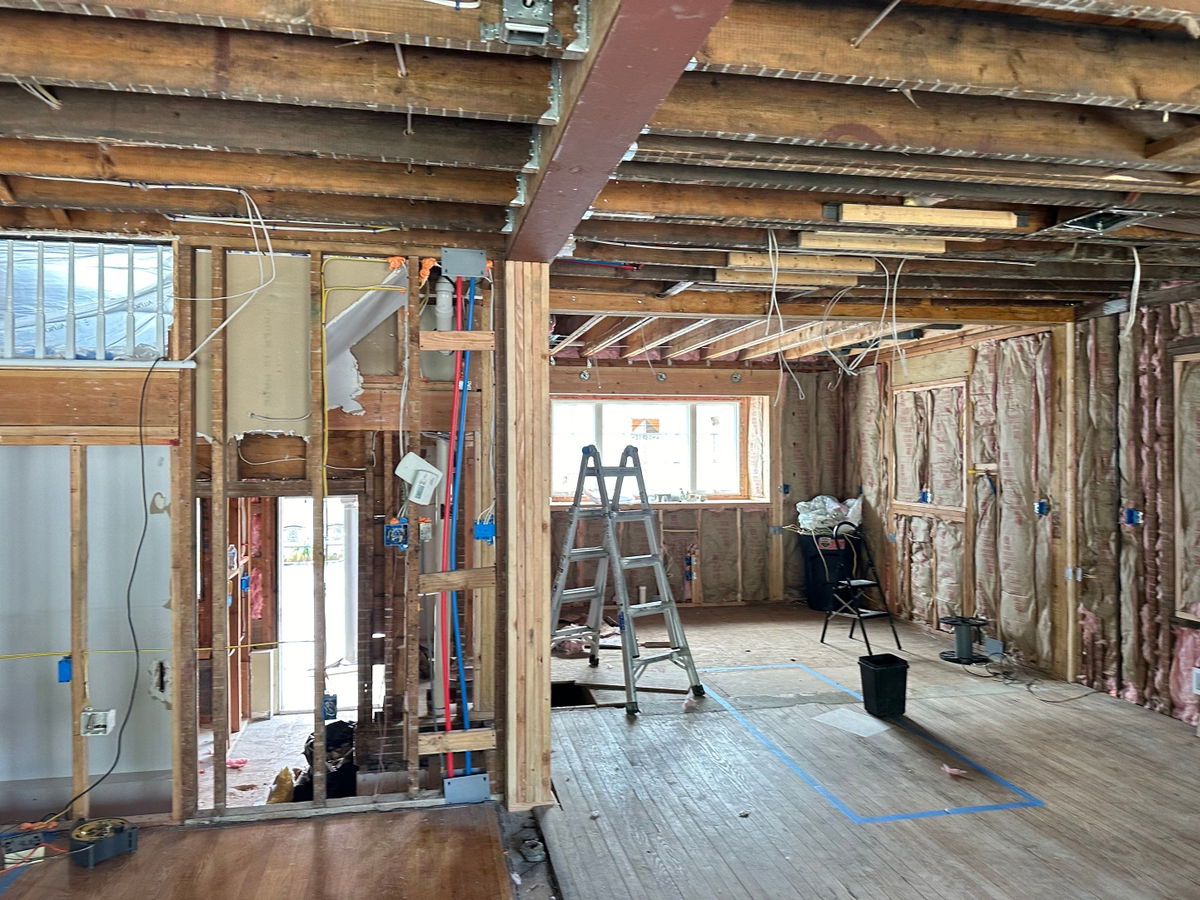 Open-concept kitchen renovation in mid-construction stage showing exposed wooden framing, beam ceiling, plumbing and electrical rough-ins, ladder, and protective plastic sheeting.