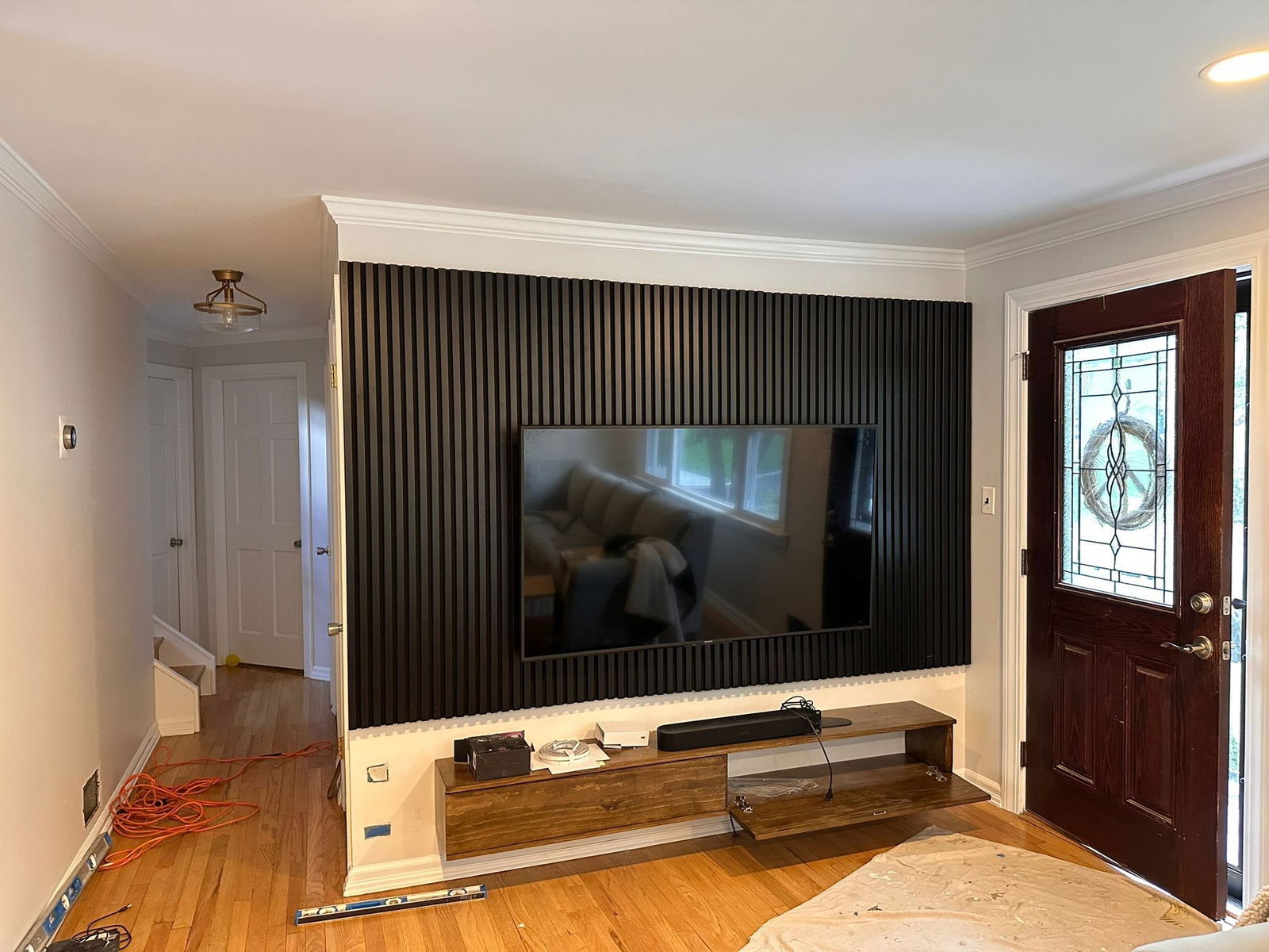 Denville interior renovation featuring a built-in media wall with black vertical slat paneling, recessed flat-screen TV mount, wooden base cabinet with open shelving, white crown molding, hardwood flooring, and dark wood entry door with le…