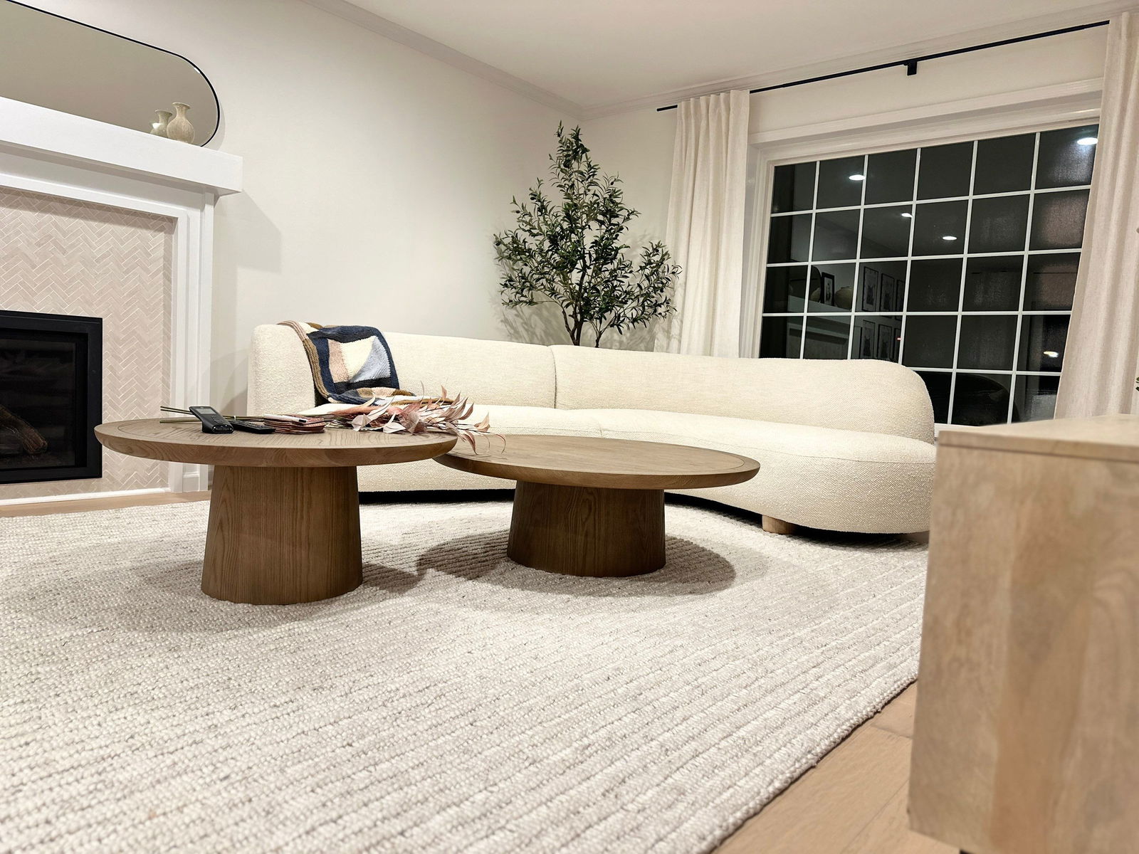 Modern living room with cream curved sofa, two wooden oval coffee tables, neutral area rug, herringbone tile fireplace surround, and black grid-pattern glass doors with white curtains.