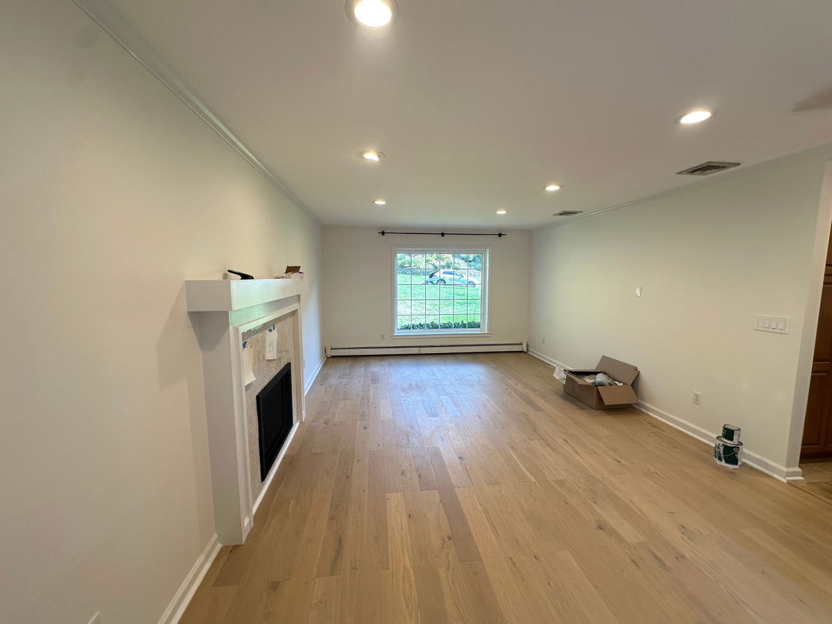 Open living room with light oak hardwood flooring, white drywall, recessed ceiling lights, a white fireplace surround on the left, and a large gridded window with black frame on the right wall.