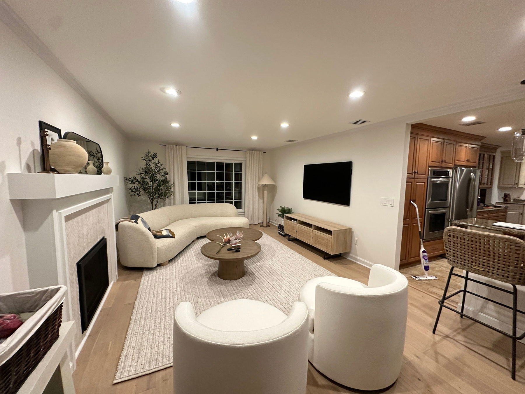 Open-concept living room with curved cream upholstered sofa, two round cream chairs, wood coffee table, mounted flat-screen TV above wood media console, white fireplace mantel with shelf styling, hardwood floors, neutral area rug, and kitc…