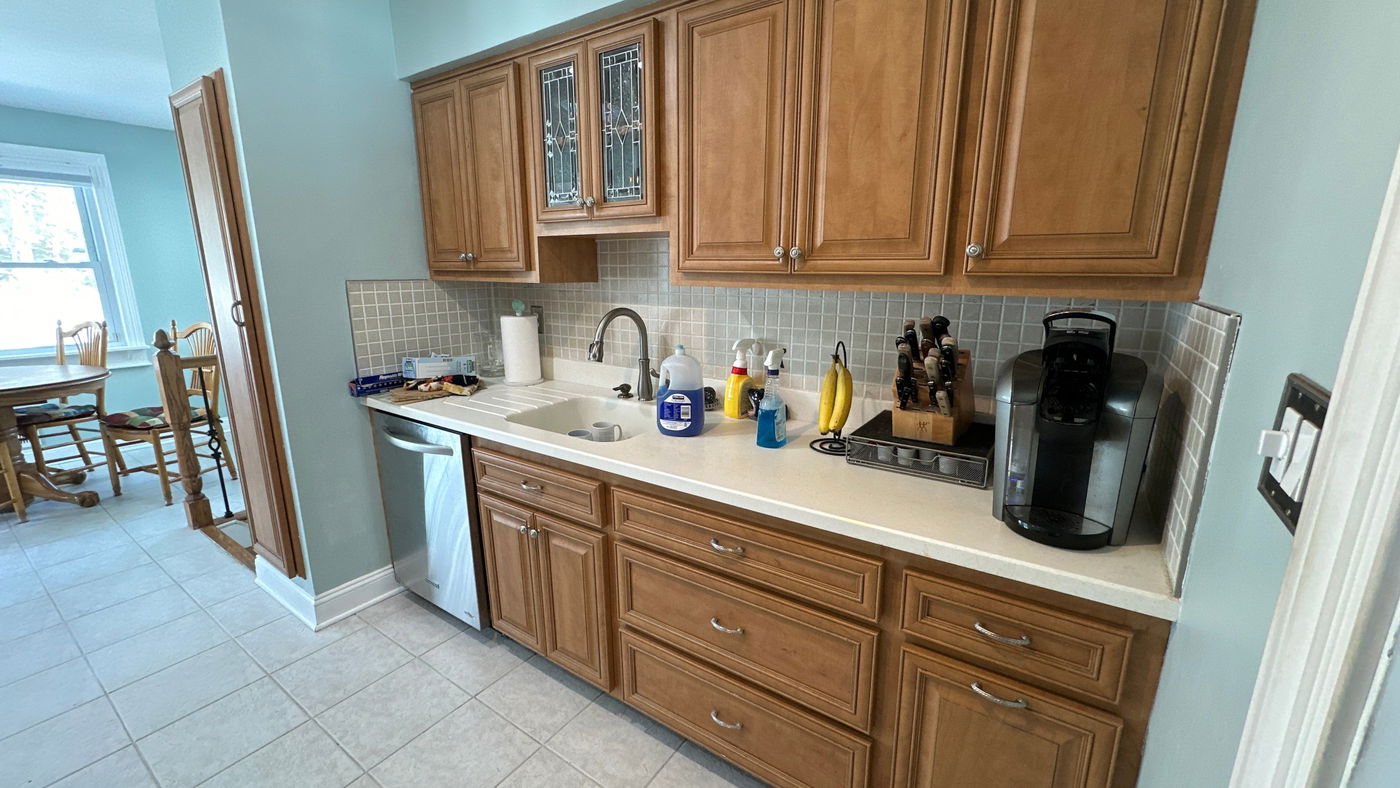 Galley kitchen with medium-tone wood cabinets, white countertop, gray subway tile backsplash, undermount sink with pull-down faucet, light blue walls, and gray ceramic tile flooring.