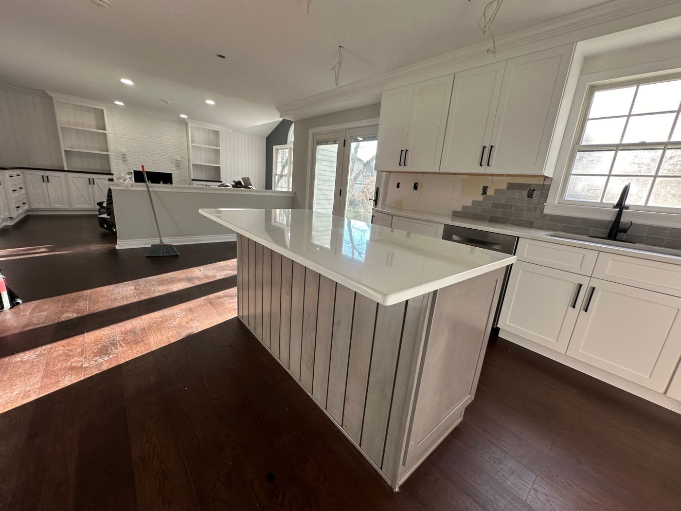 Open-concept kitchen with white shaker cabinets, white quartz waterfall island, gray subway tile backsplash, dark wood flooring, and large windows overlooking exterior.
