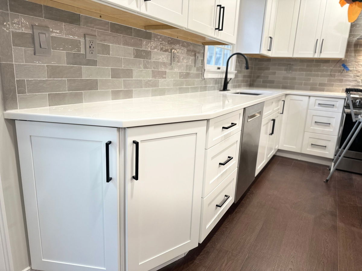 White shaker cabinets with black hardware, white quartz countertop, gray subway tile backsplash, black matte faucet, and dark wood-look flooring in a galley kitchen layout.