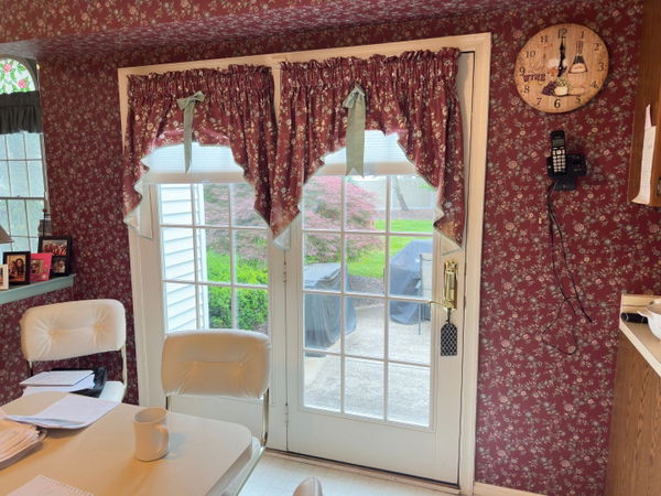Burgundy floral wallpapered room with French doors, burgundy damask curtains with sage ties, white wood trim, and natural light from multi-pane glass doors overlooking green landscape.