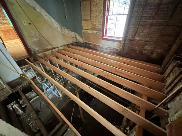 Exposed wooden floor joists and subfloor framing in gutted kitchen space with brick walls, peeling plaster, and a burgundy-trimmed window.