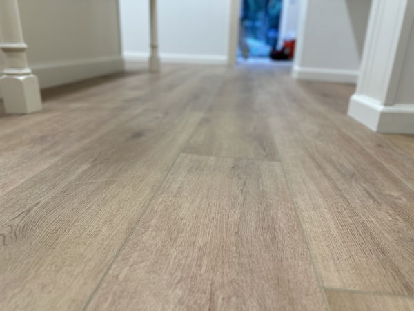 Wide-plank wood-look flooring in light gray-tan tones with white baseboards and trim, extending through an open space with large windows in background.