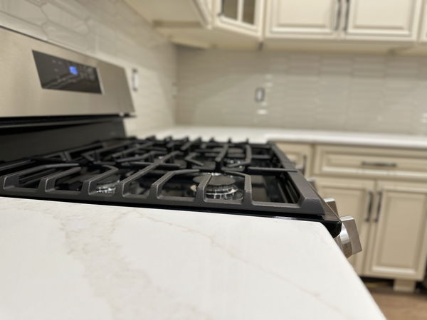 Black gas cooktop with multiple burners on white marble countertop, cream shaker cabinets and stainless steel appliances in background.