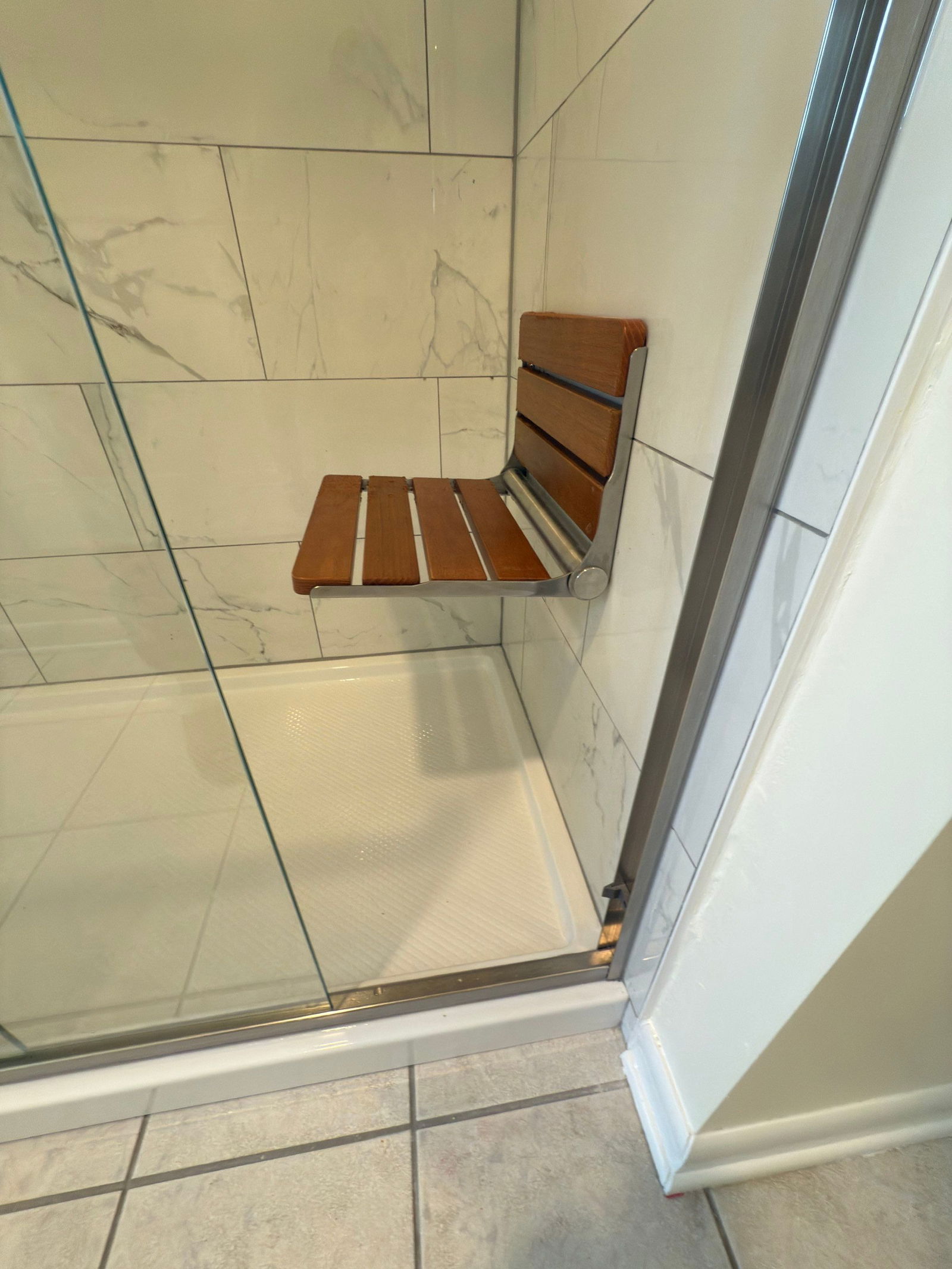 Walk-in shower with cream marble-look tile walls, linear drain system, white base pan, and teak wood bench seat with chrome hardware.
