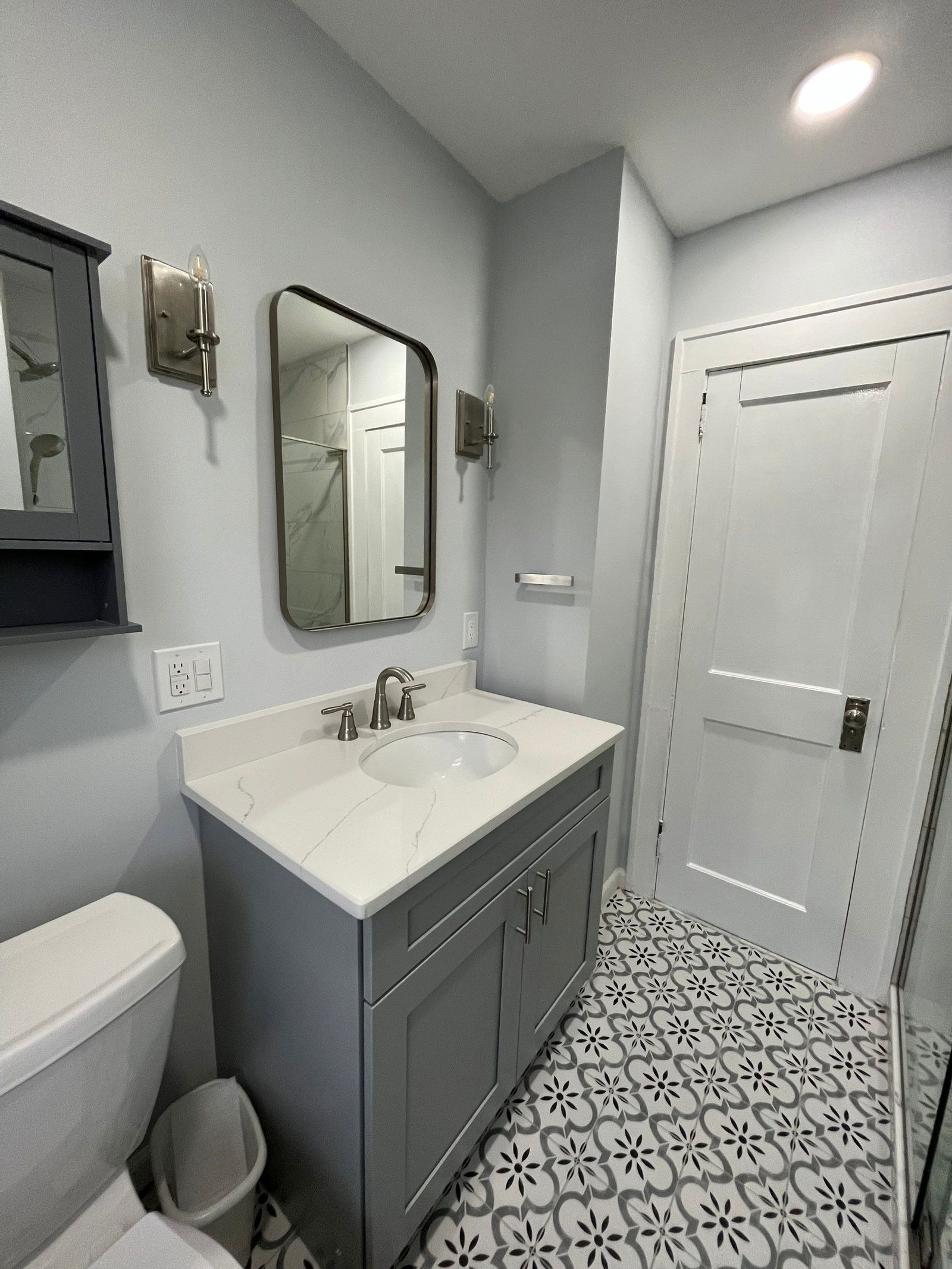 Modern bathroom with gray vanity, white quartz countertop, round undermount sink, black-framed mirror, brushed nickel faucet, gray subway tile walls, patterned floor tile with geometric design, white shaker doors, and recessed ceiling ligh…