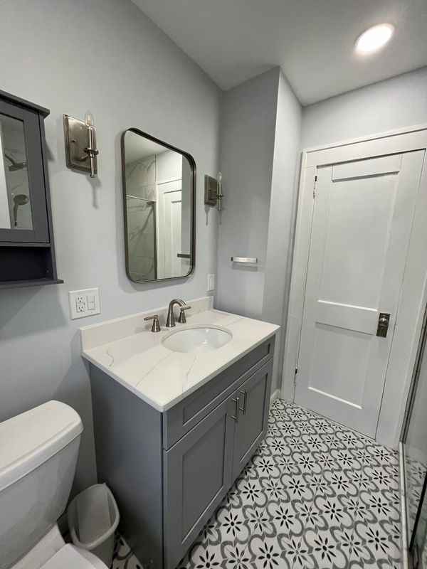 Modern bathroom with gray vanity, white quartz countertop, round undermount sink, black-framed mirror, brushed nickel faucet, gray subway tile walls, patterned floor tile with geometric design, white shaker doors, and recessed ceiling ligh…
