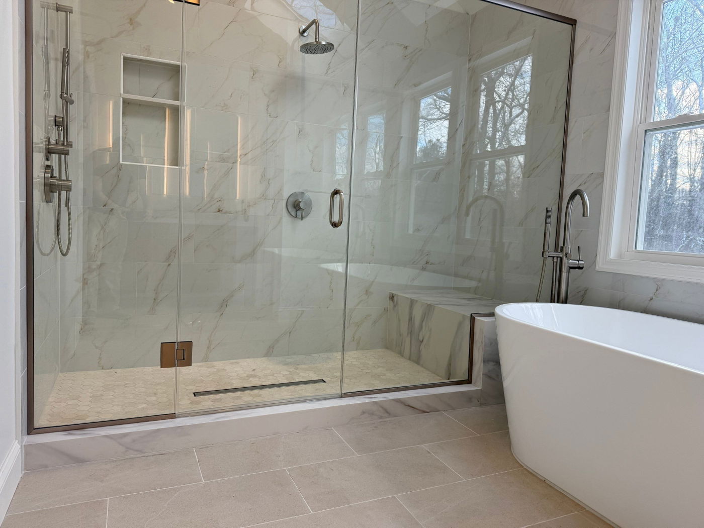 Spacious master bathroom with marble-tile walk-in shower, frameless glass enclosure, rainfall showerhead, white freestanding soaking tub, neutral tile flooring, and windows overlooking landscaping.